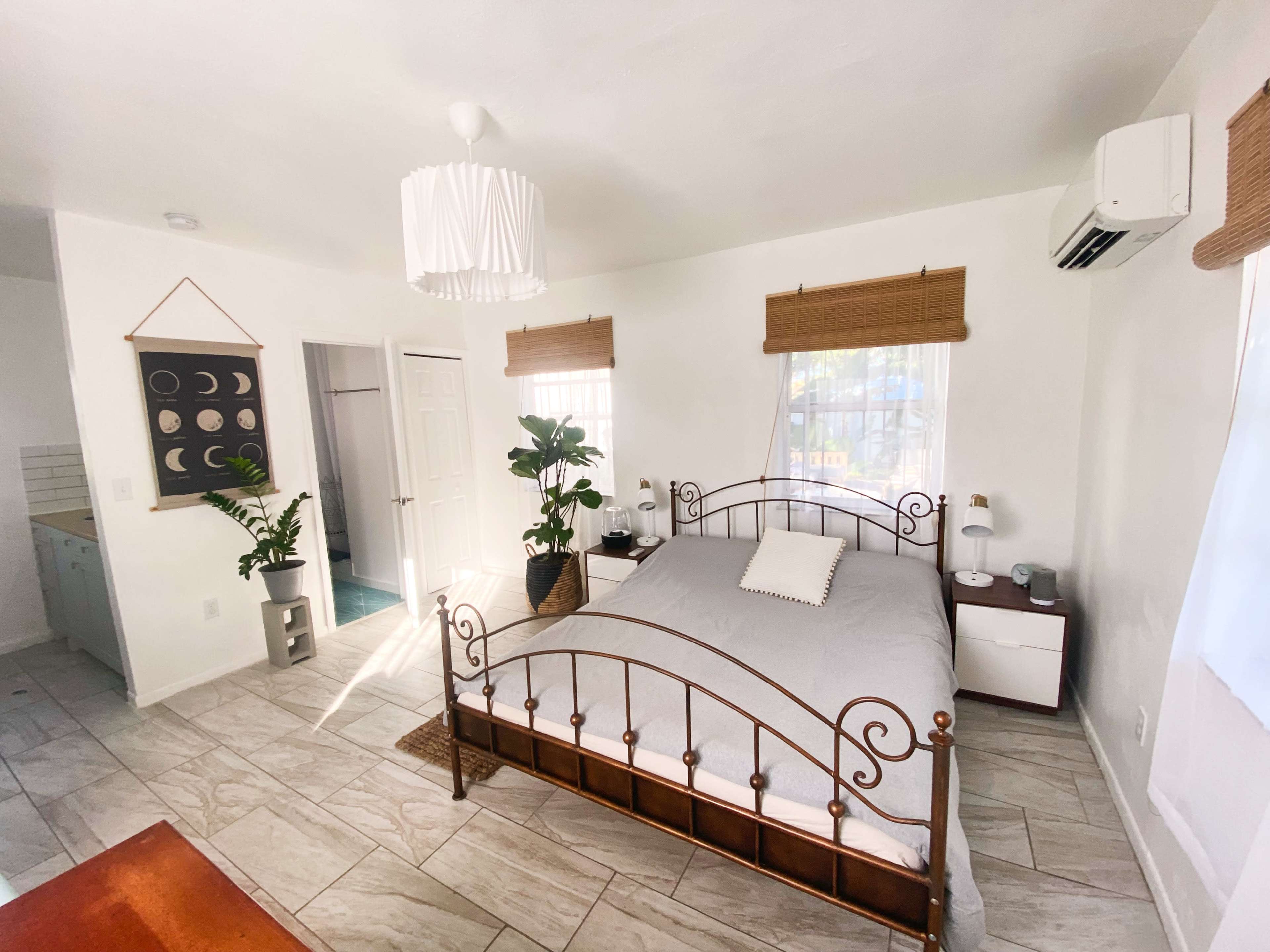 The image shows a well-lit bedroom featuring a metal bed frame with a light gray quilt, wooden nightstands, and indoor plants, alongside an open door leading to a bathroom area.