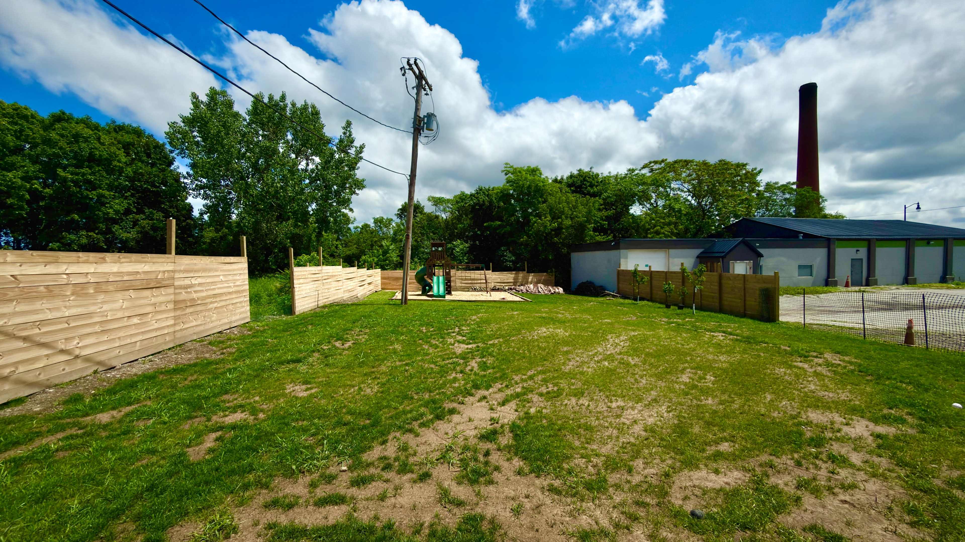 The image shows a grassy area enclosed by wooden panels, with a power pole and a industrial building in the background under a partly cloudy sky.