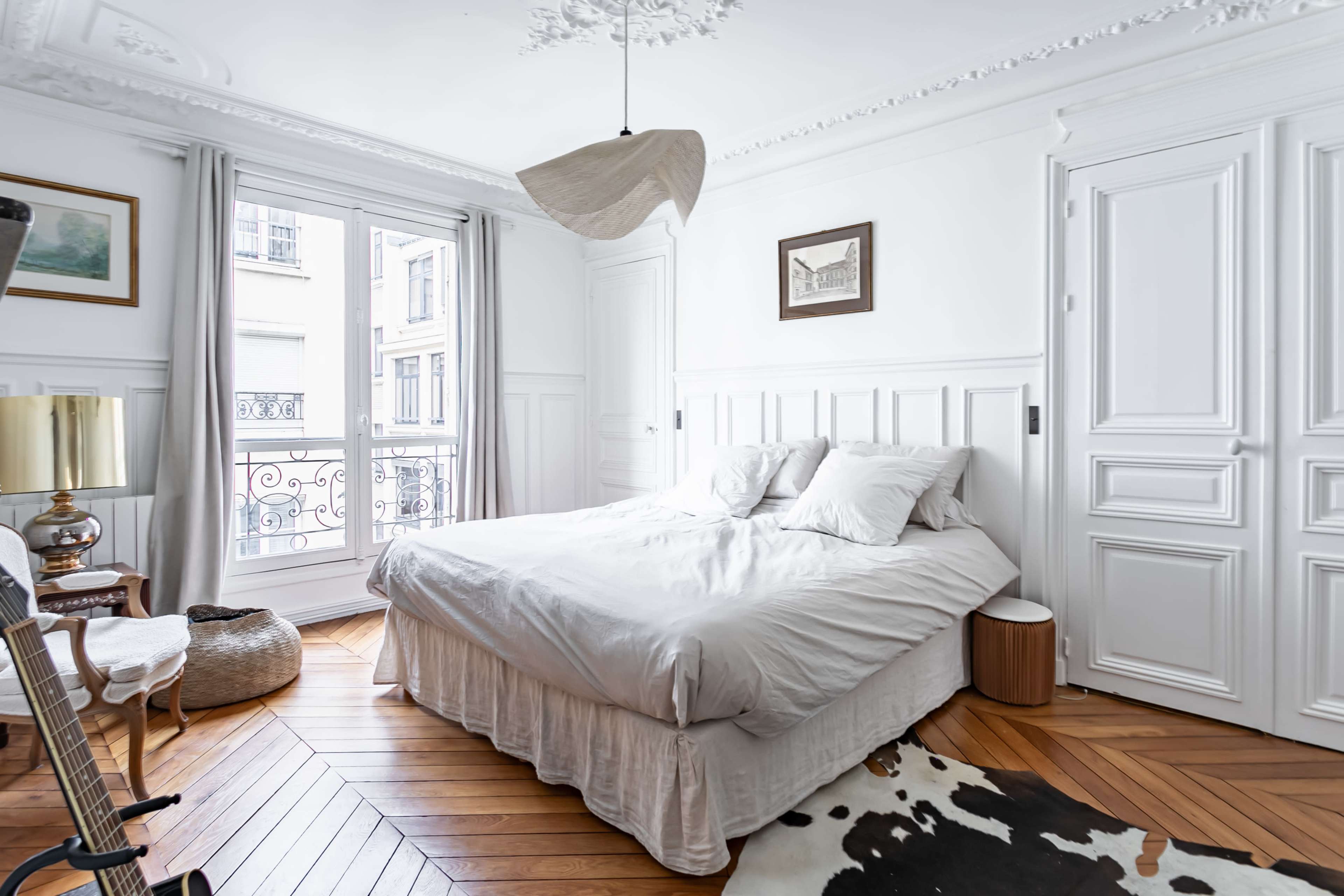 A bright bedroom with a double bed, two windows, and wooden flooring, featuring a guitar and a decorative rug.