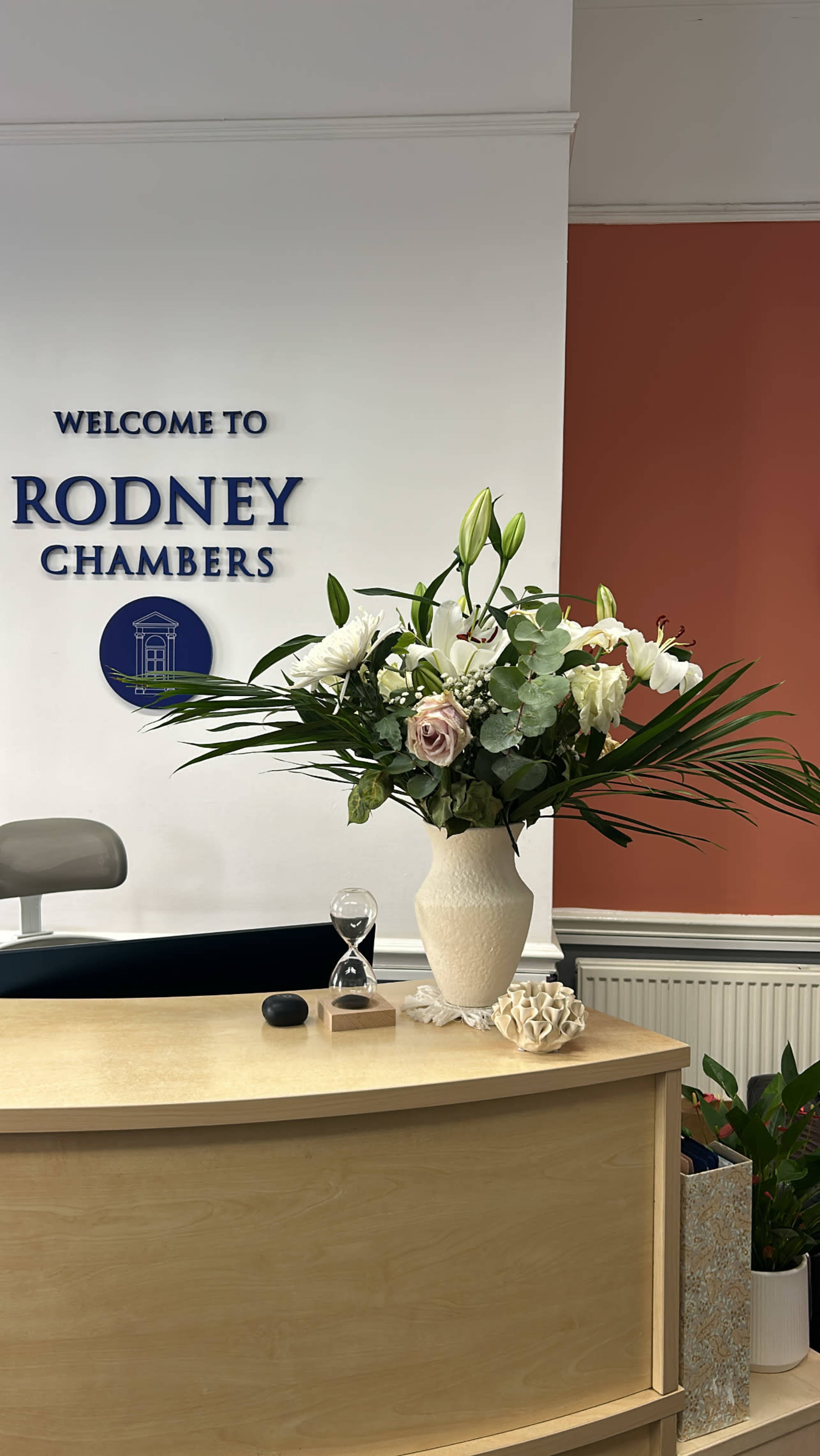 The reception area features a decorative arrangement of flowers in a vase on a wooden desk, with a welcome sign for "Rodney Chambers" on the wall behind.
