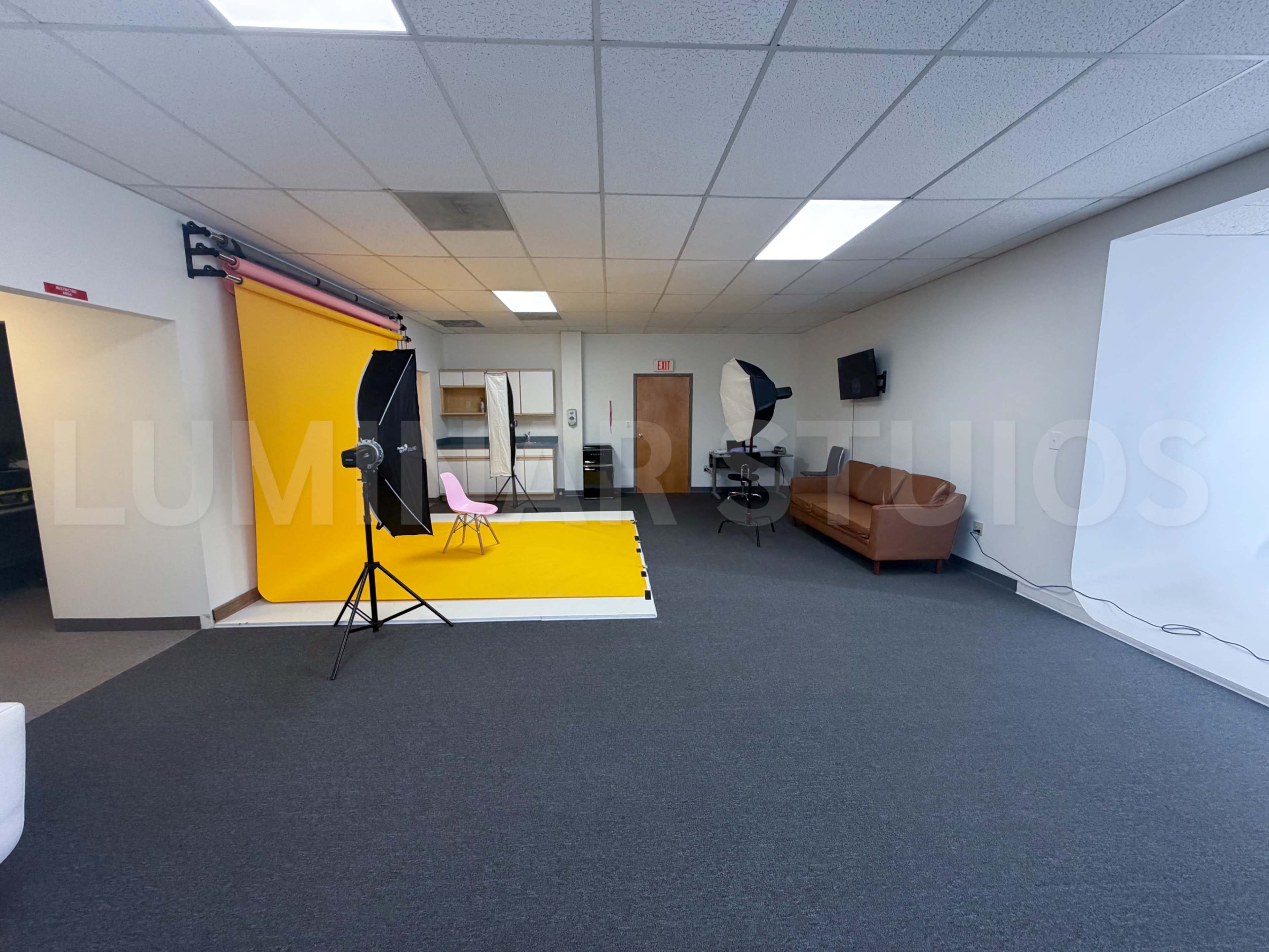 The image shows a photography studio with a yellow backdrop, lighting equipment, and a brown couch against a plain wall.