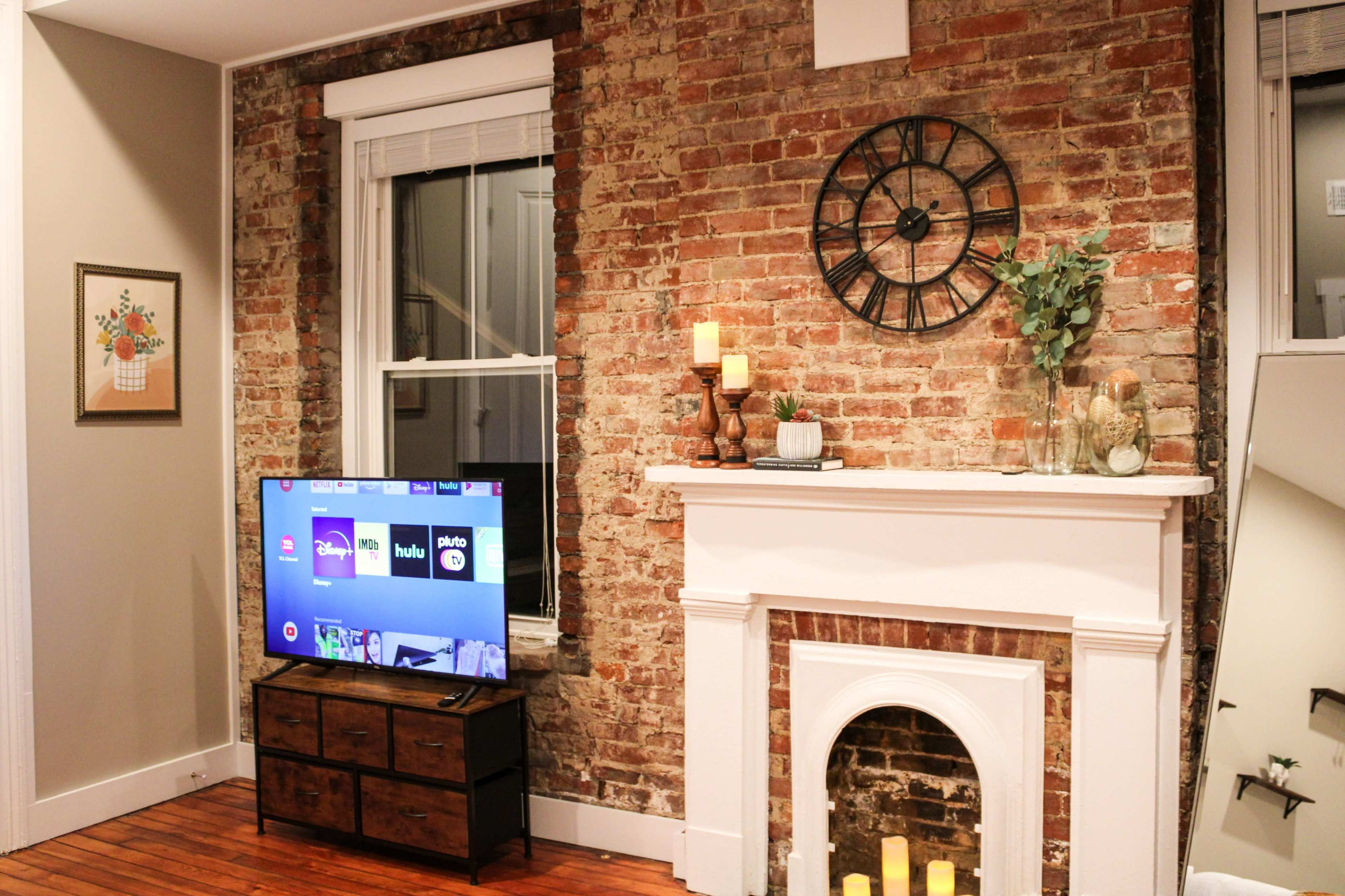 A living room with a brick wall, a television on a wooden stand, and a fireplace with candles and decorative items.