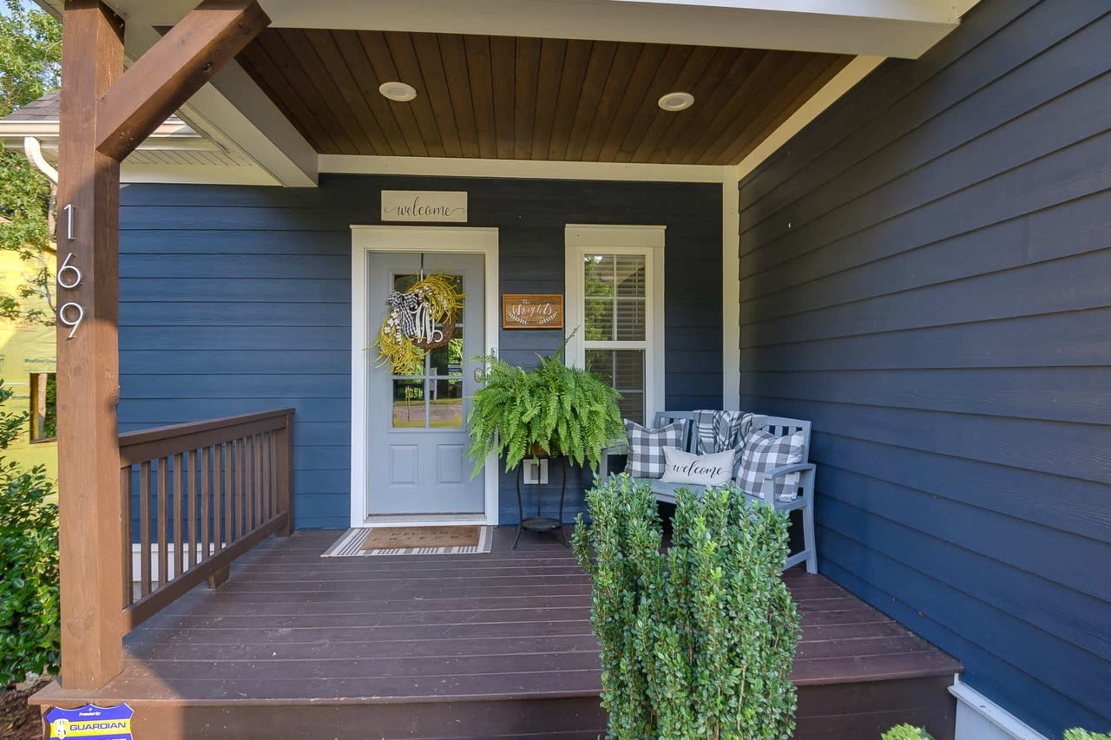 The image shows a front porch with a blue exterior, featuring a welcome sign, potted fern, checkered chairs, and decorative elements.