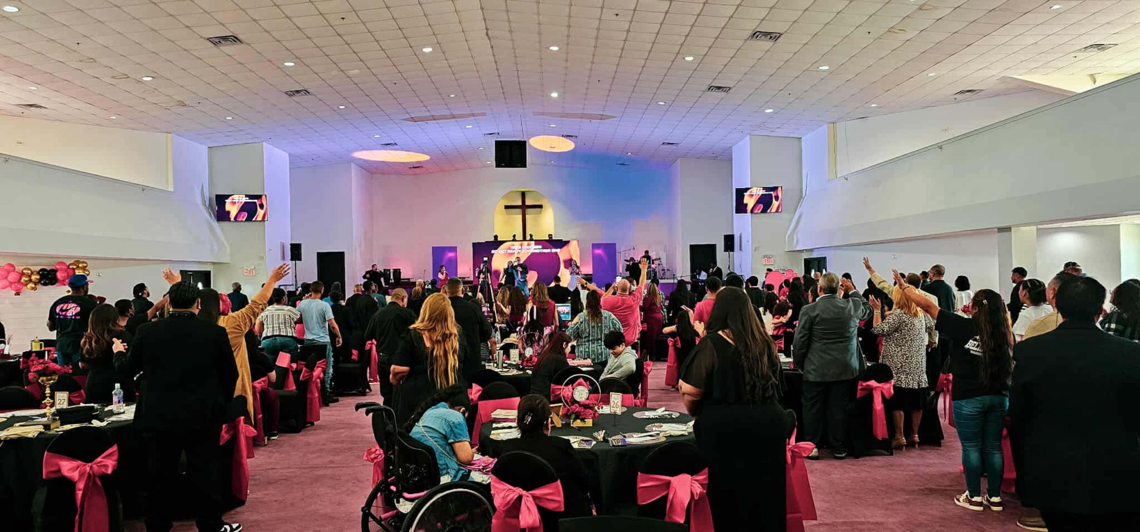 A large gathering of people is worshiping inside a spacious hall decorated with pink accents and a cross prominently displayed in the background.