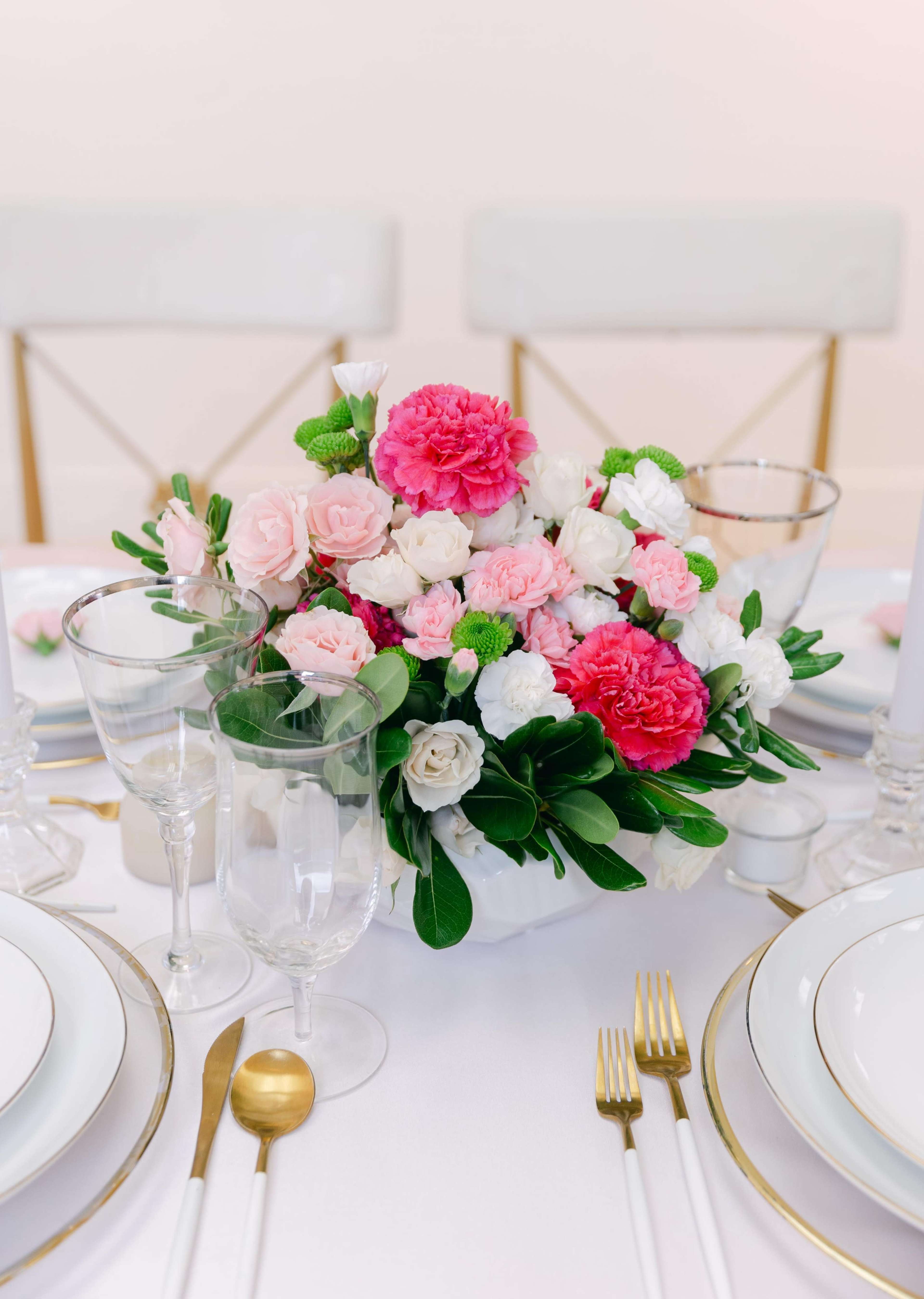 A vibrant floral centerpiece featuring pink, white, and green flowers is displayed on a elegantly set dining table with golden utensils and glassware.