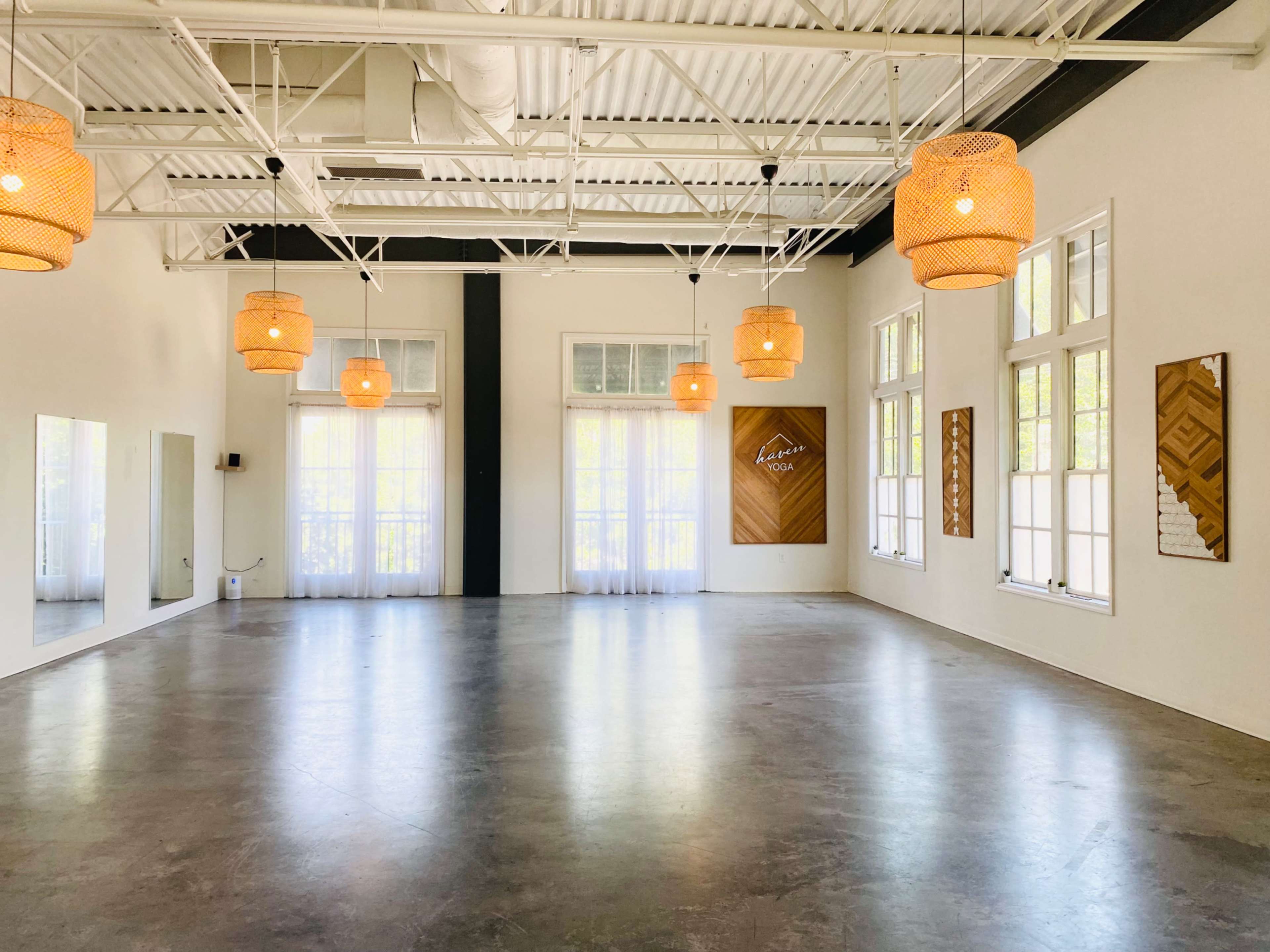 The image shows a spacious, well-lit room with wooden pendant lights, large windows, and a polished concrete floor.