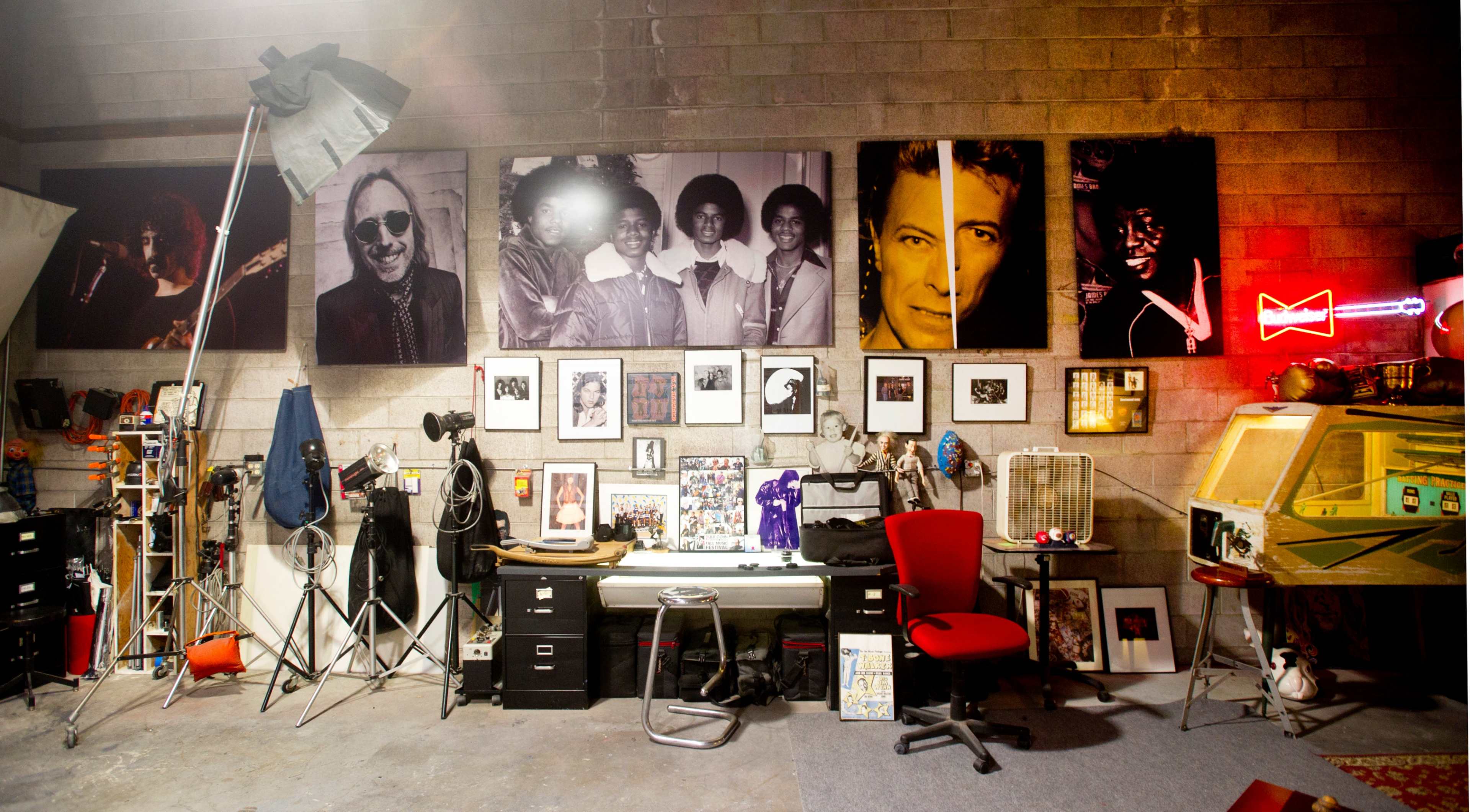 A studio space with large black-and-white photographs on the wall, various photography equipment, and a red chair in front of a desk filled with art and memorabilia.