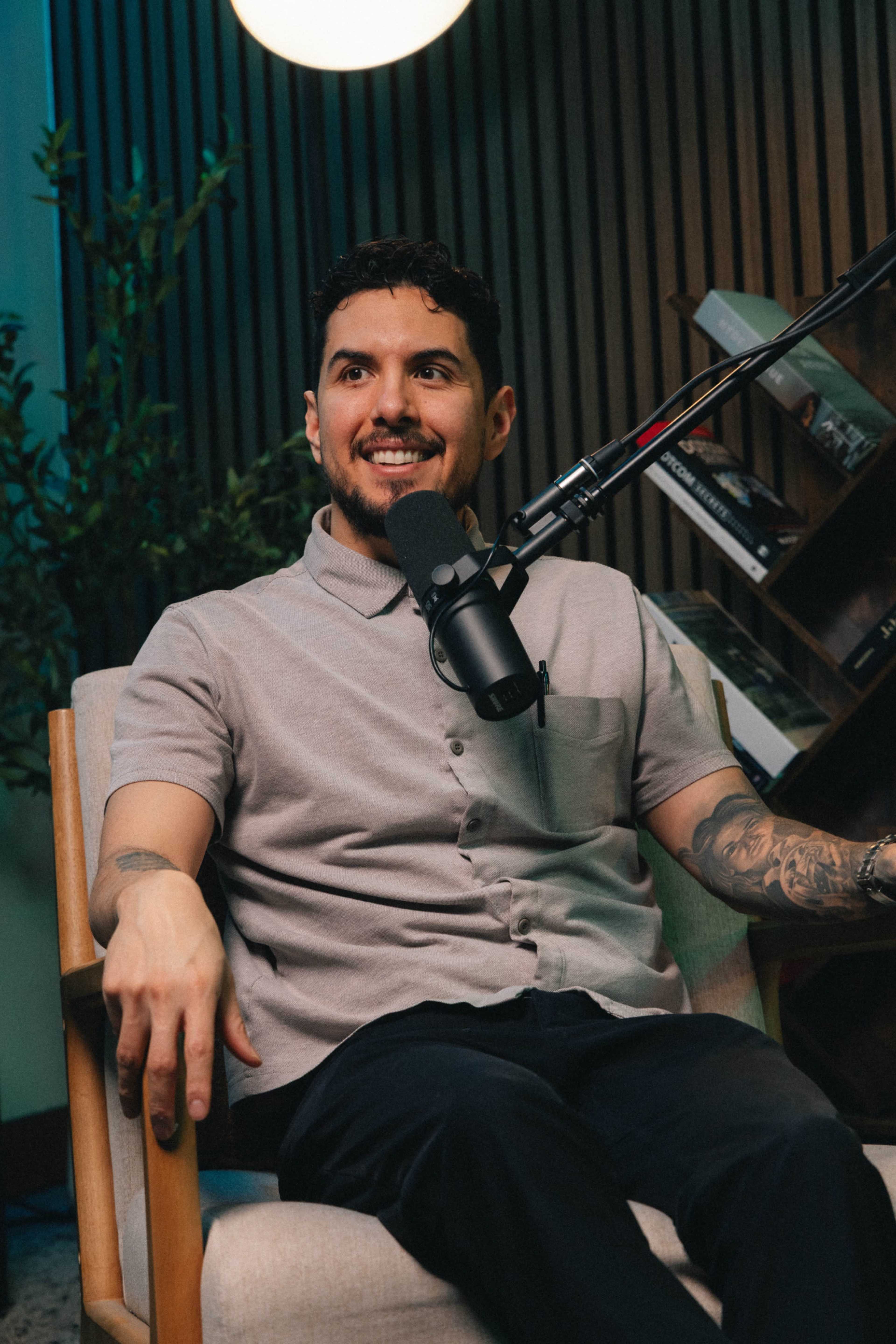 A man with short dark hair and a grey polo shirt sits in a chair, smiling while speaking into a microphone during a podcast recording.