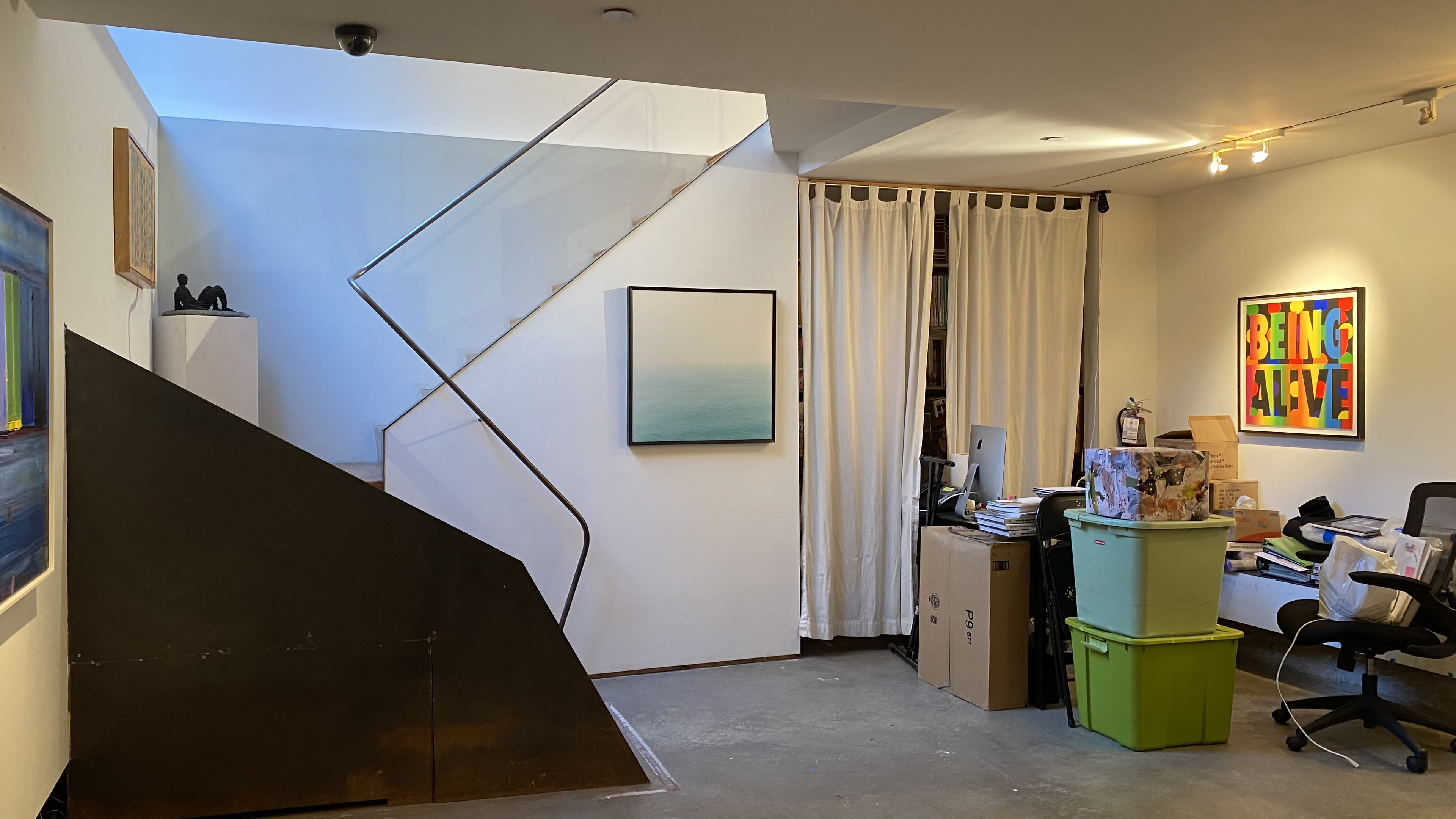 The image shows a modern interior space with a staircase leading to another level, a desk cluttered with boxes and supplies, and artwork on the walls.