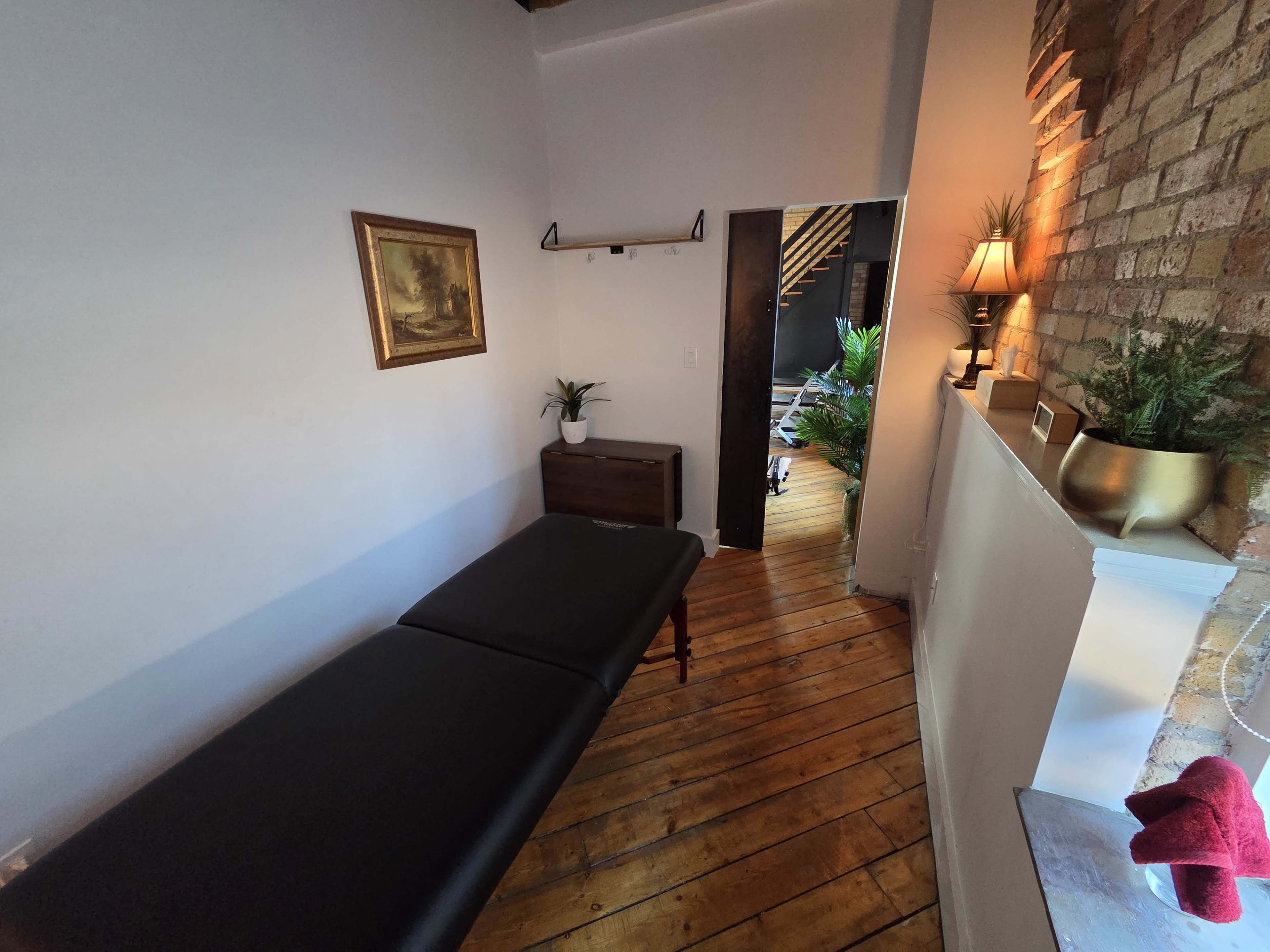 The image shows a calming treatment room with a massage table, wooden flooring, a small decorative chest, and plants in the background.