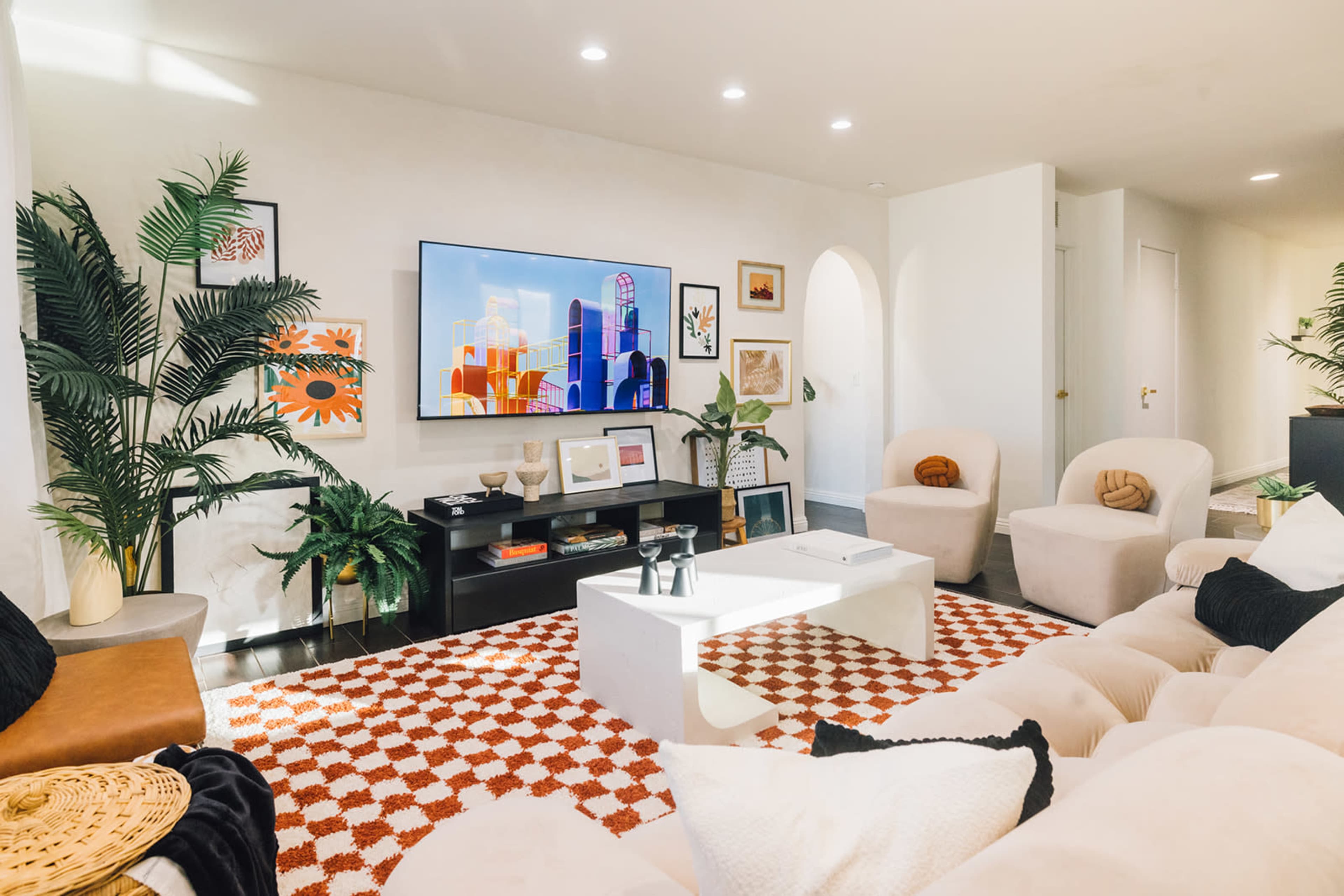 A contemporary living room features a large television mounted on the wall, a beige sofa, a coffee table, and decorative plants along with various artworks and a patterned rug.