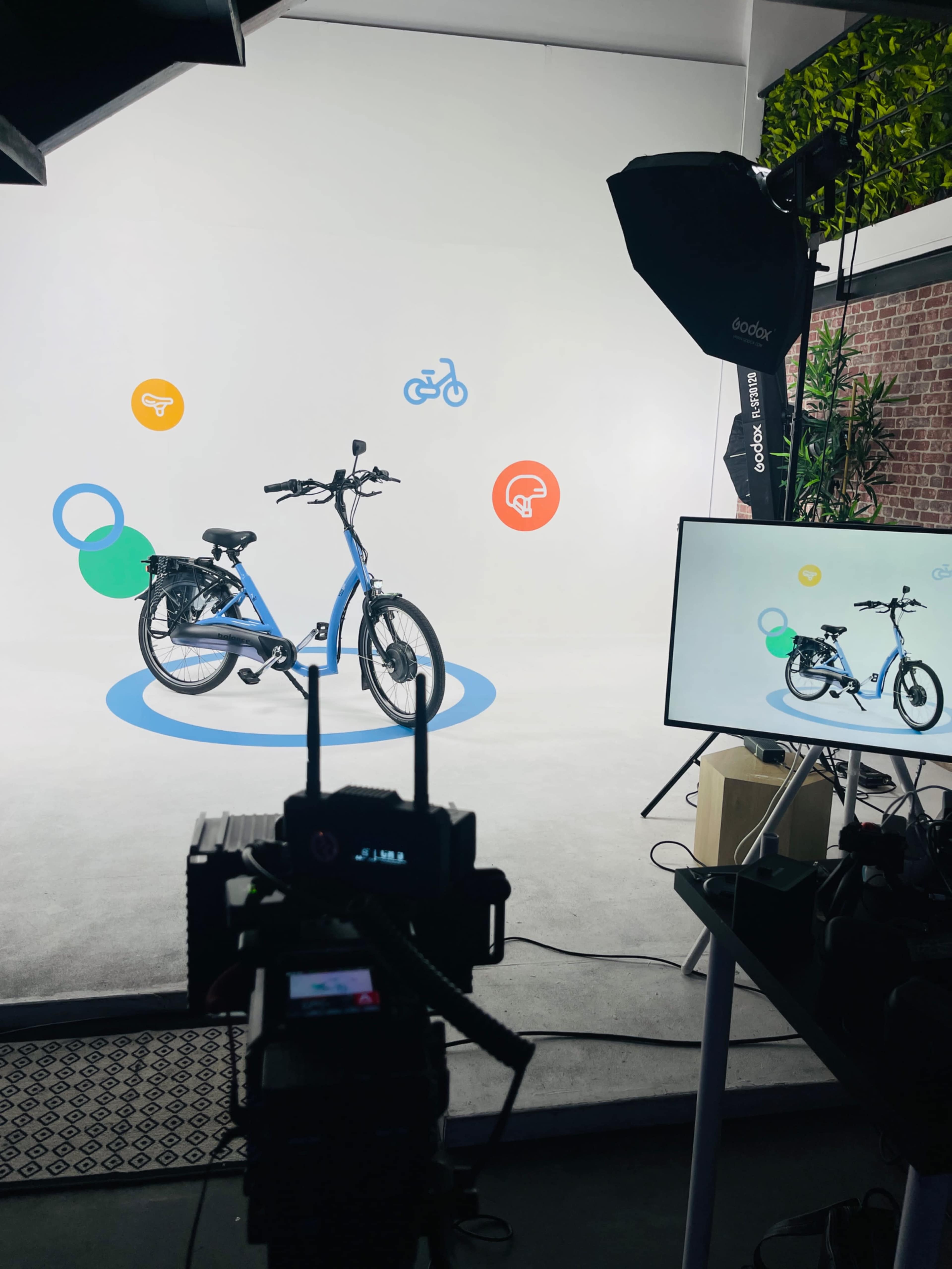 A blue bicycle is positioned on a plain background with colorful graphic icons surrounding it, while a camera and a monitor are set up to capture the scene.