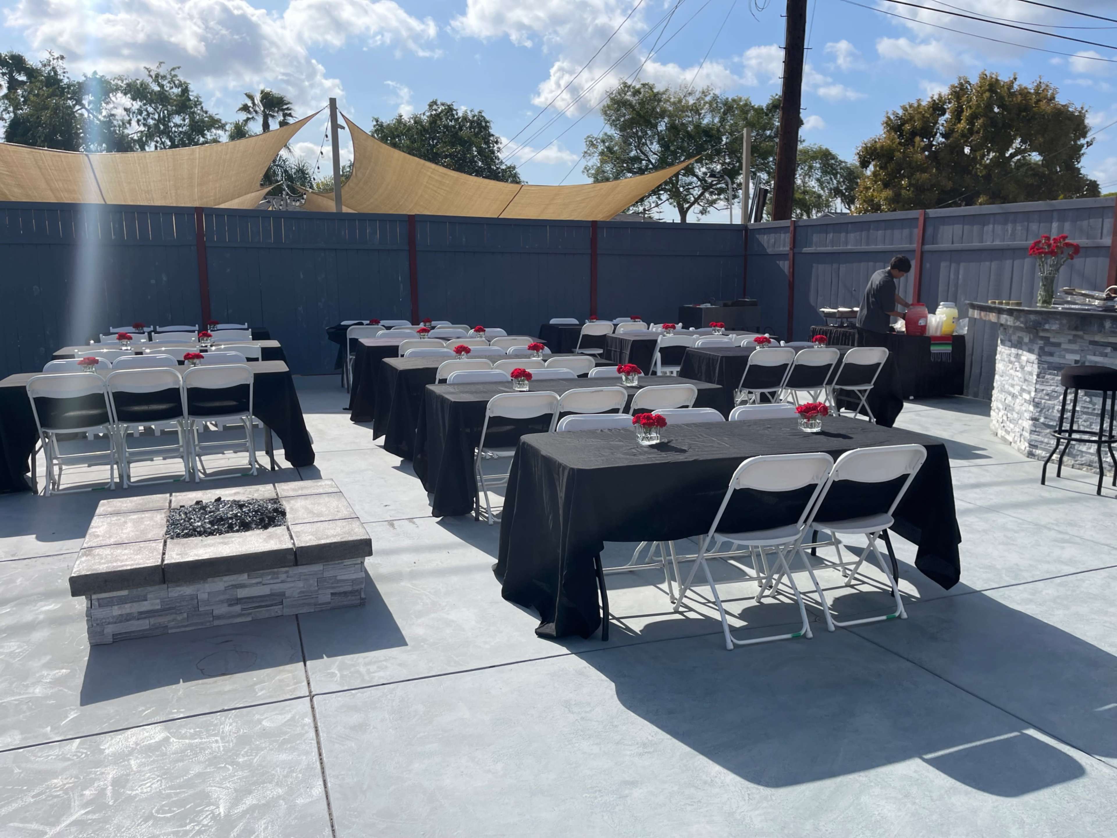 The image shows an outdoor event space with multiple tables covered in black tablecloths, each adorned with a small flower centerpiece, and a service area with refreshments in the background.