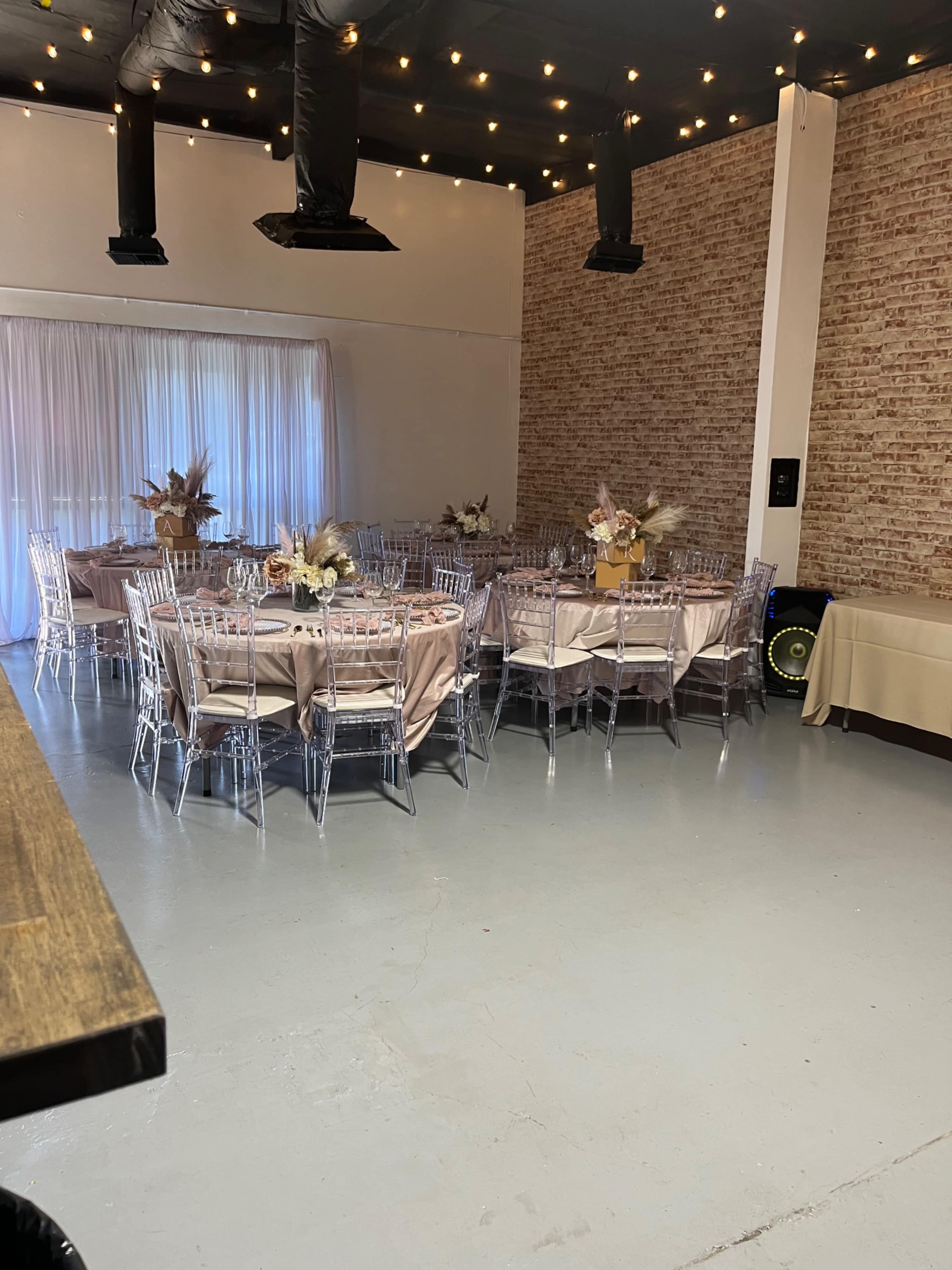 A dimly lit event space with several round tables set for a gathering, each adorned with light-colored tablecloths and centerpieces, and decorated with string lights overhead.