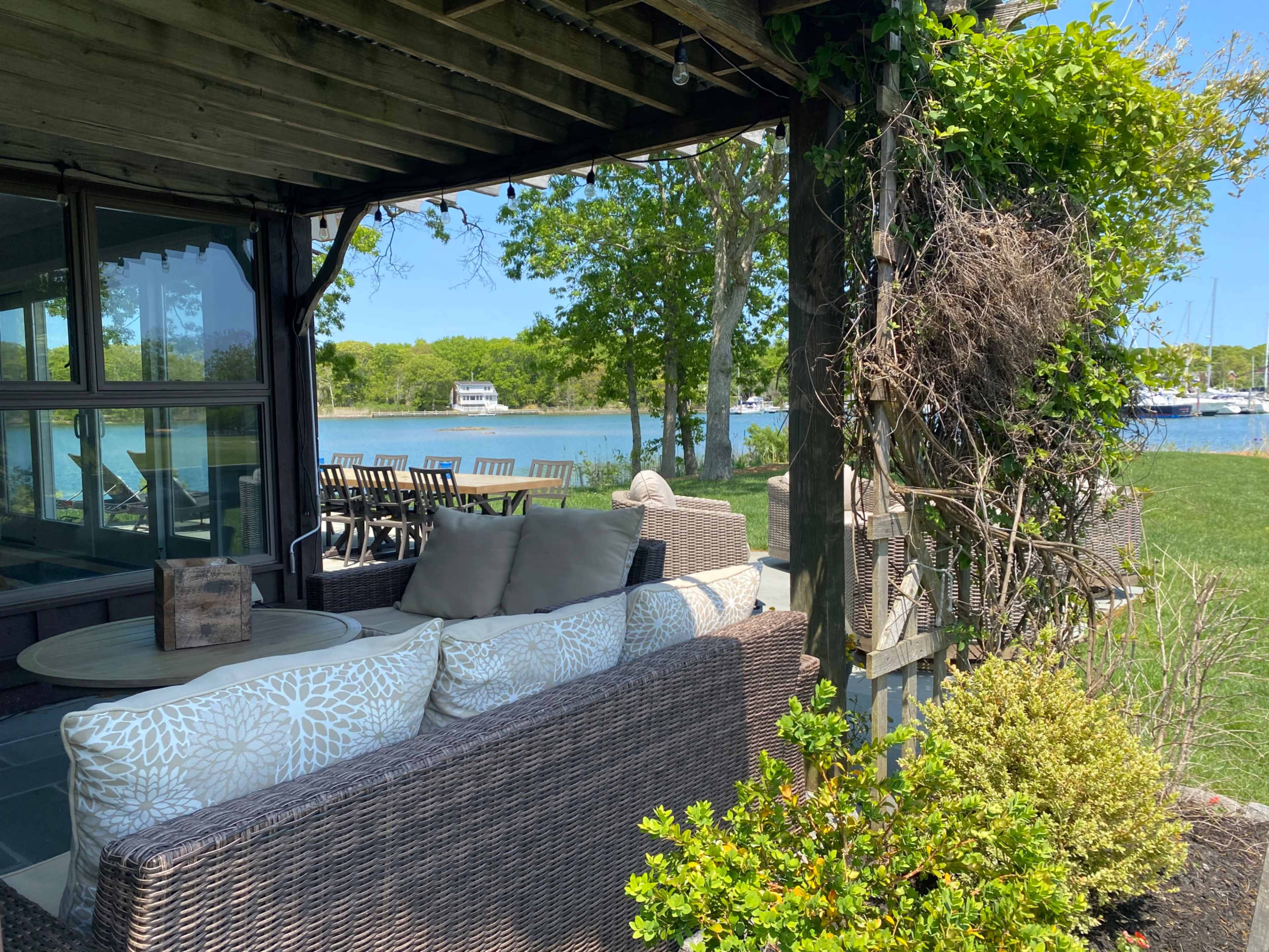A covered patio with wicker furniture overlooks a calm waterfront with boats in the distance.