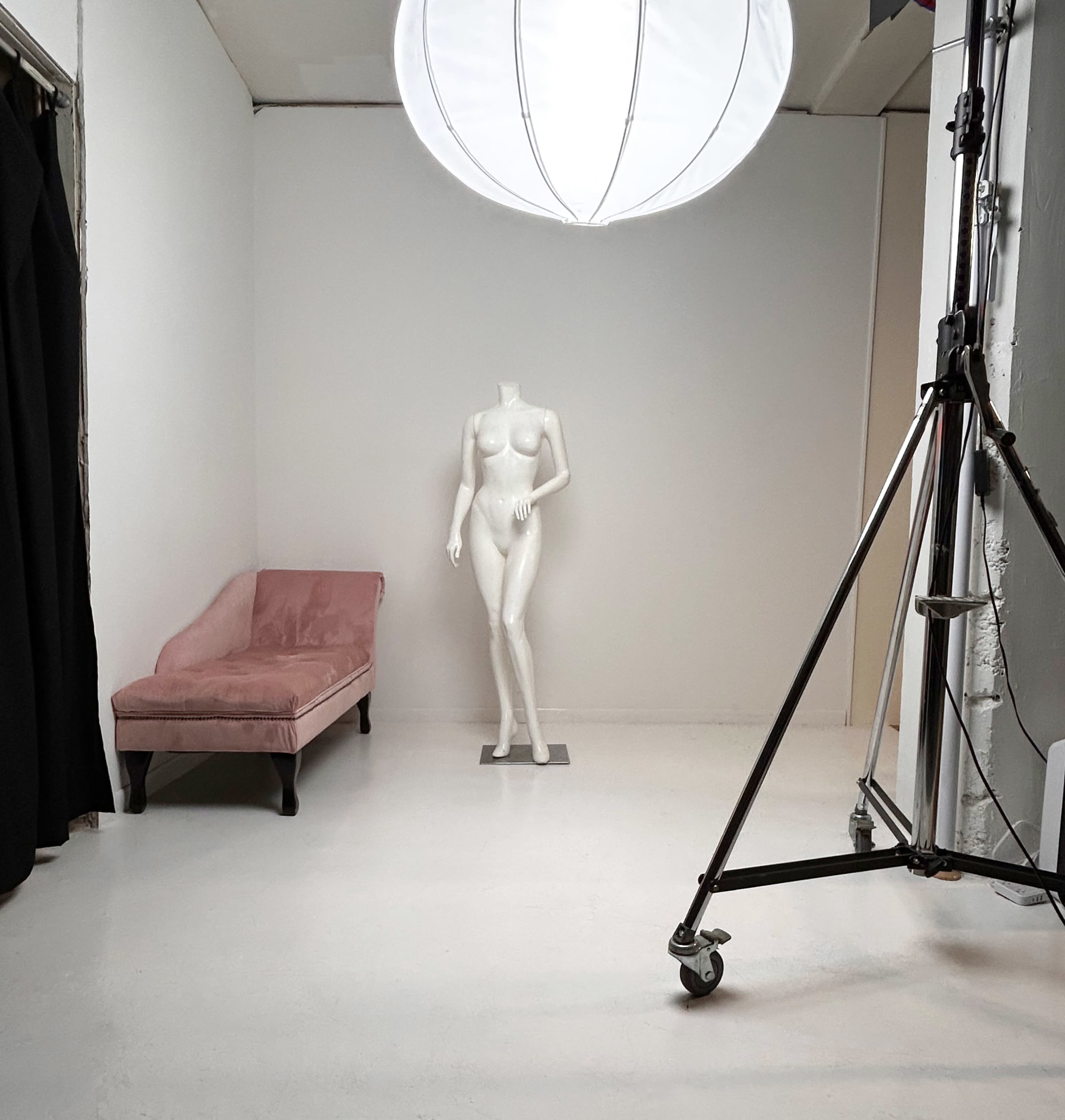 The image shows a minimalist studio setting with a mannequin standing beside a pink velvet sofa and a large light fixture overhead.