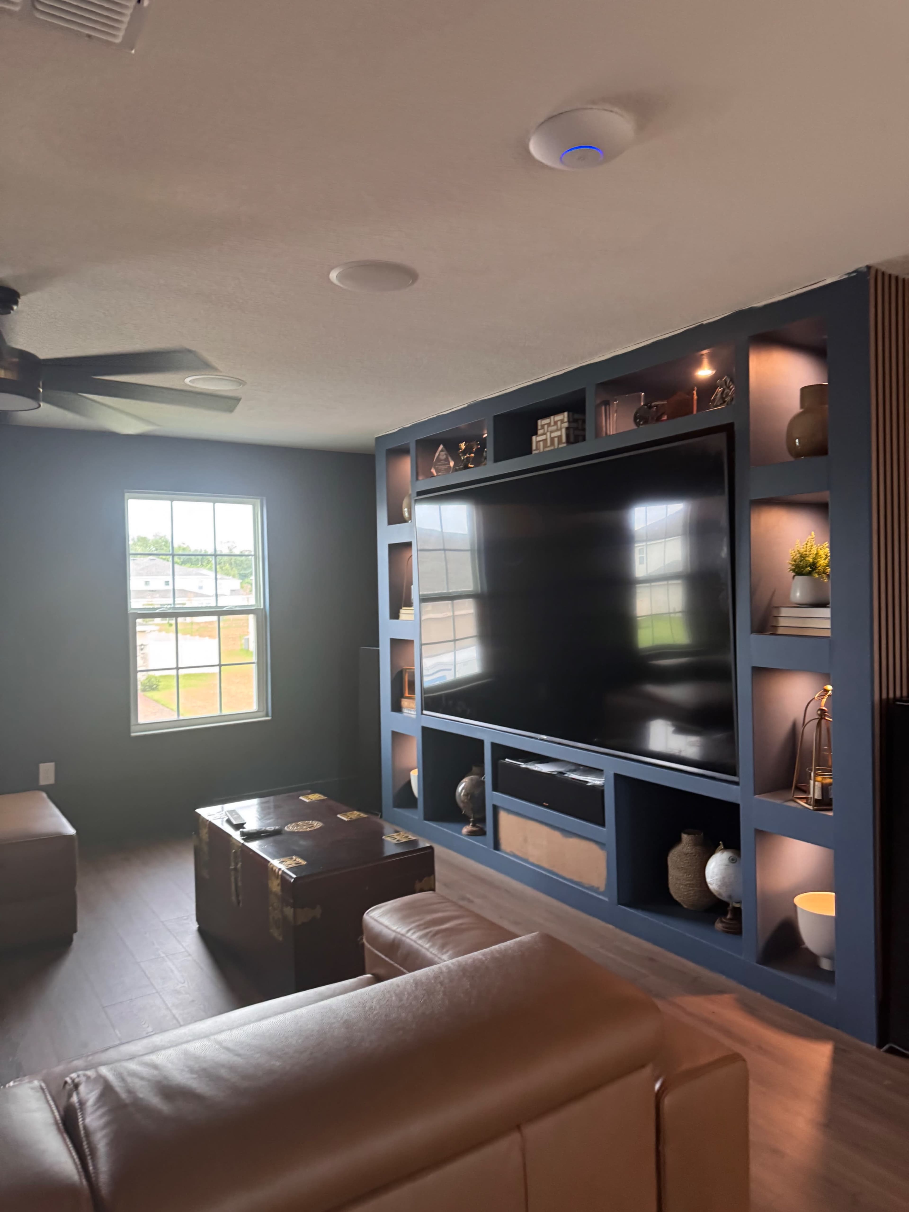 The image shows a modern living room with a large wall-mounted television, built-in shelves displaying decorative items, and a window providing natural light.