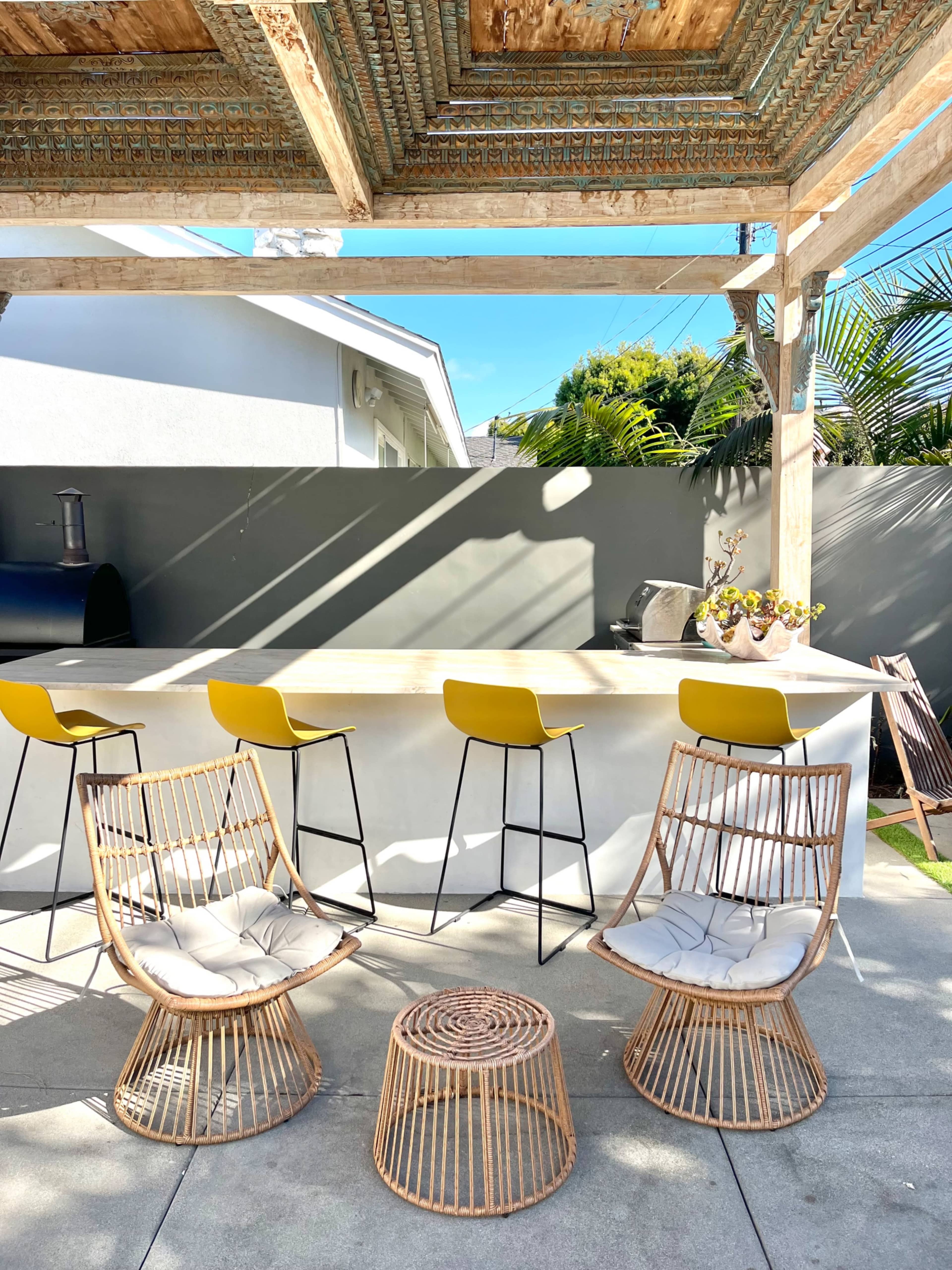 An outdoor bar area features four rattan chairs, a round coffee table, and a counter with yellow stools under a pergola.