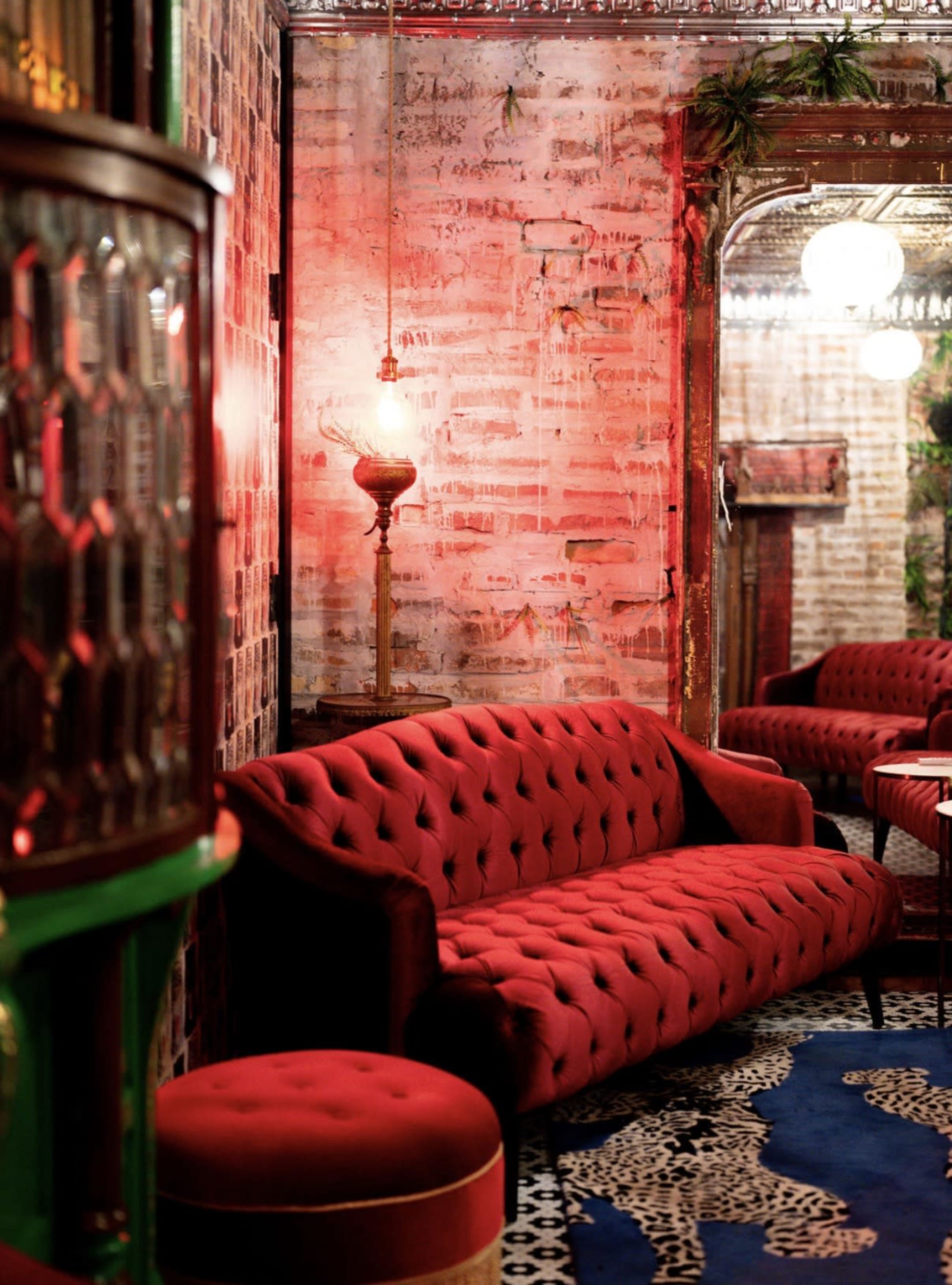 The image shows a vintage-style lounge area with a red tufted sofa, a matching ottoman, and patterned carpeting, all set against a background of exposed brick walls and ornate mirrors.