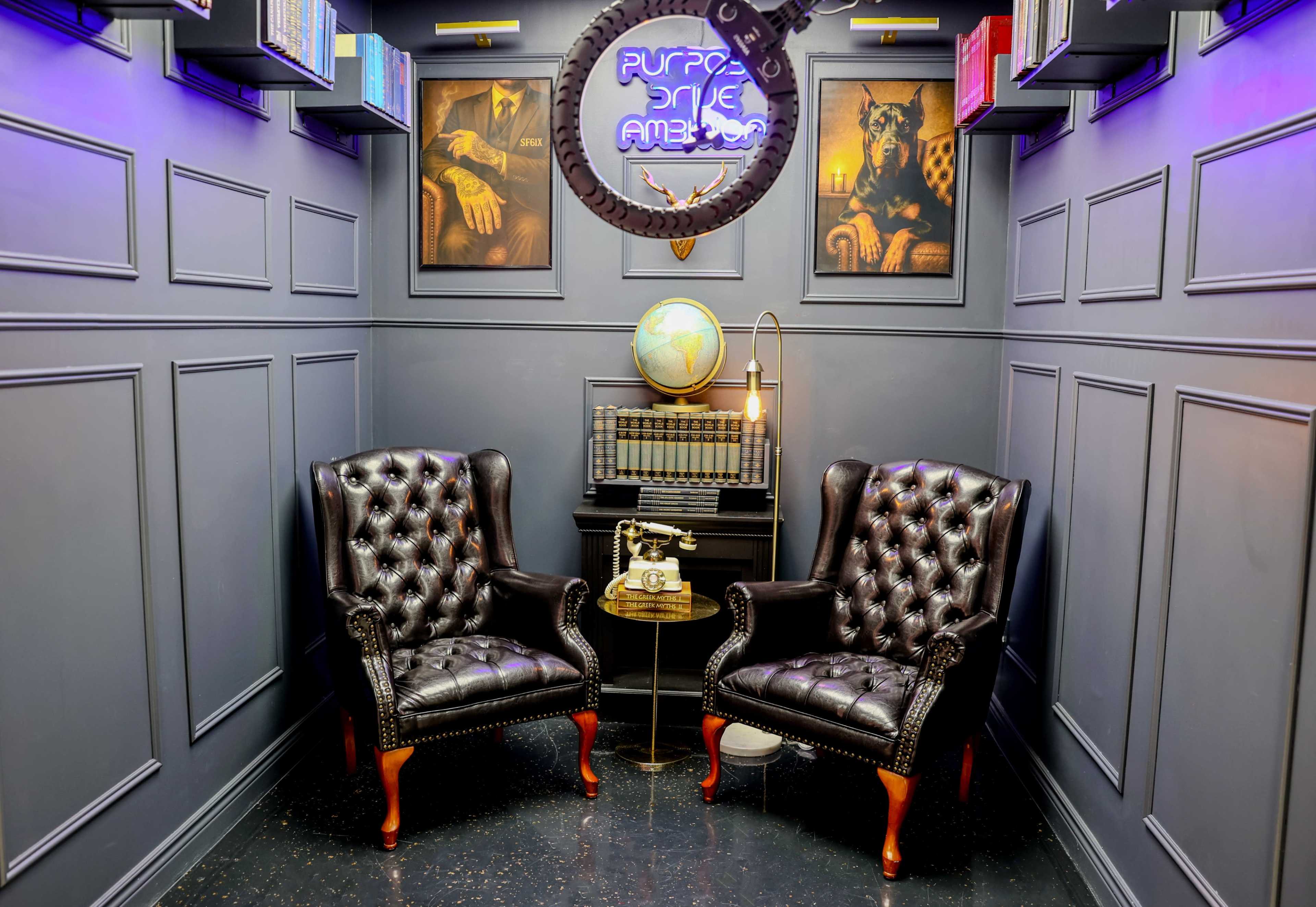 The image shows a cozy reading nook featuring two tufted leather armchairs, a small wooden table, and framed artwork on the walls, illuminated by soft lighting.