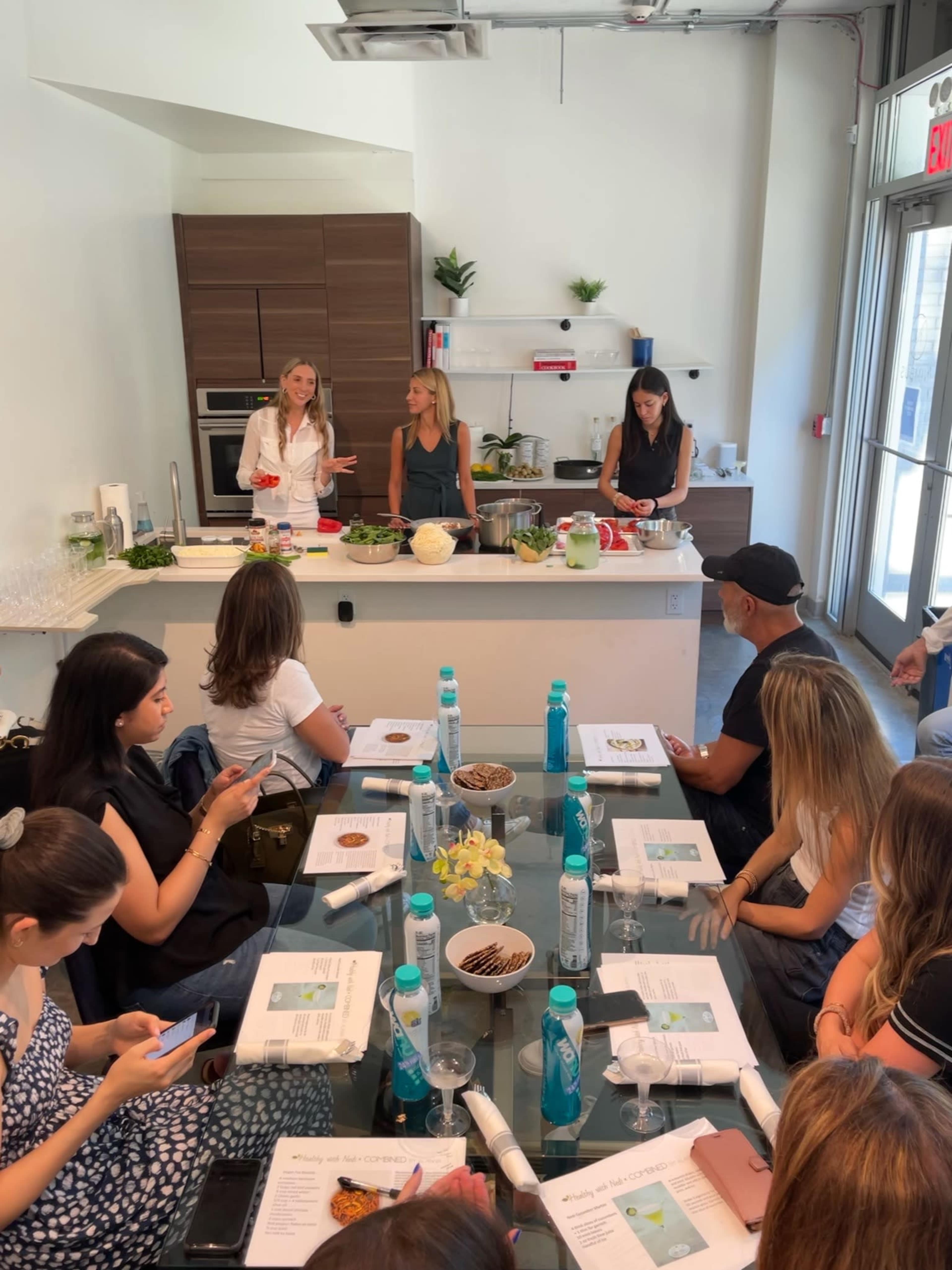 A group of people sit at a table in a kitchen, watching two individuals demonstrate a cooking lesson while another prepares ingredients.