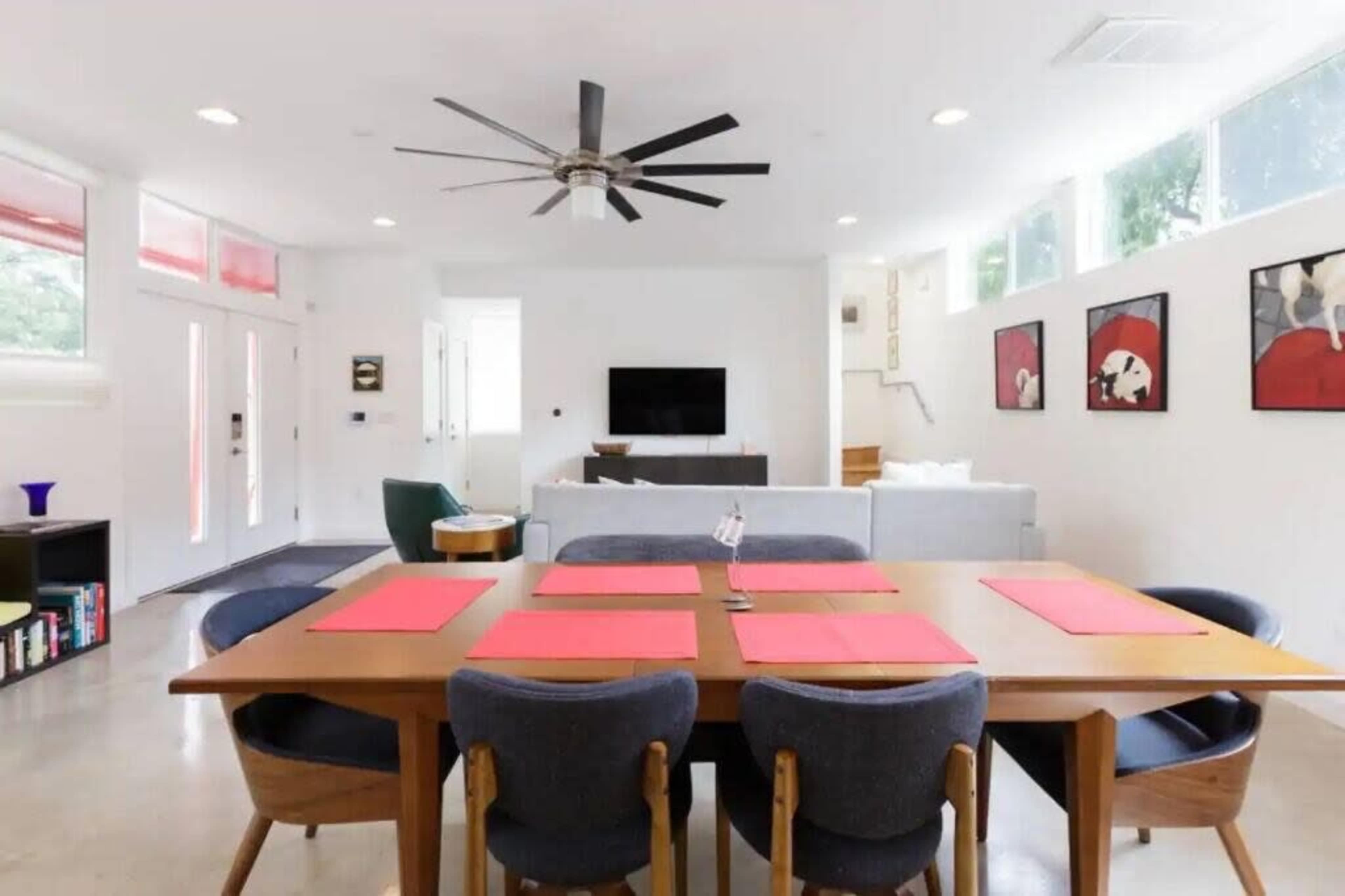 A dining table with red placemats is set in a bright, modern living space featuring a fan, a sofa, and wall art.