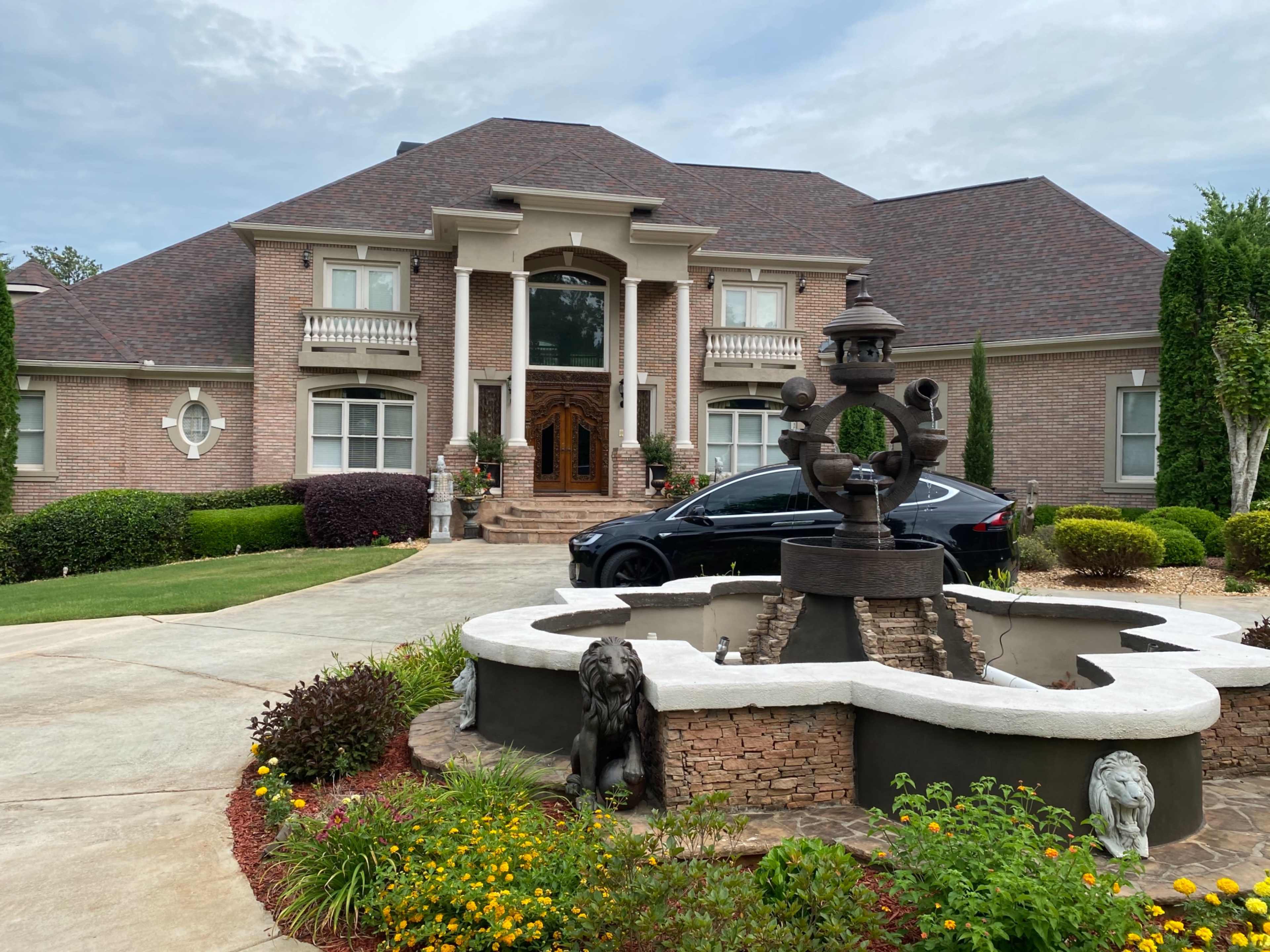 A large brick house with a fountain and sculptures in the front yard, alongside a driveway leading to a black car.