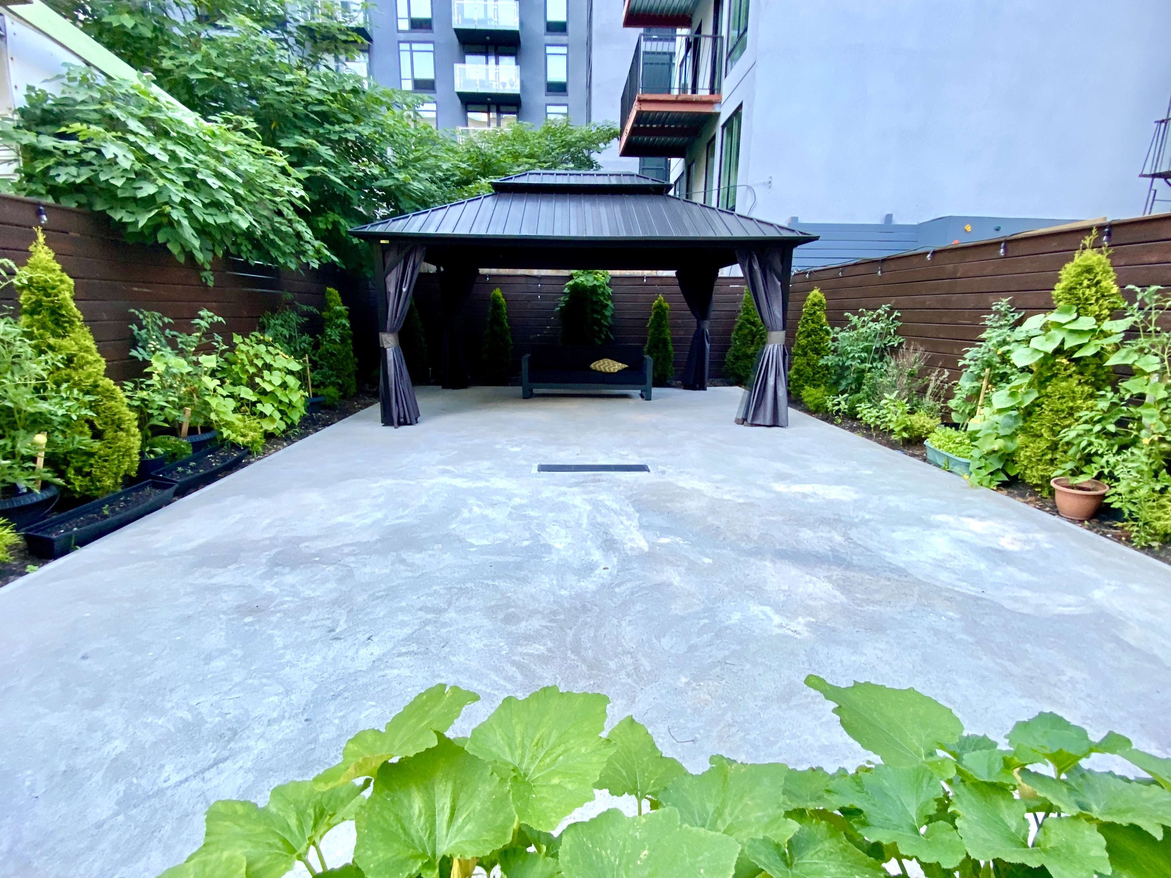 A concrete outdoor area featuring a gazebo surrounded by neatly arranged greenery, including small shrubs and potted plants.