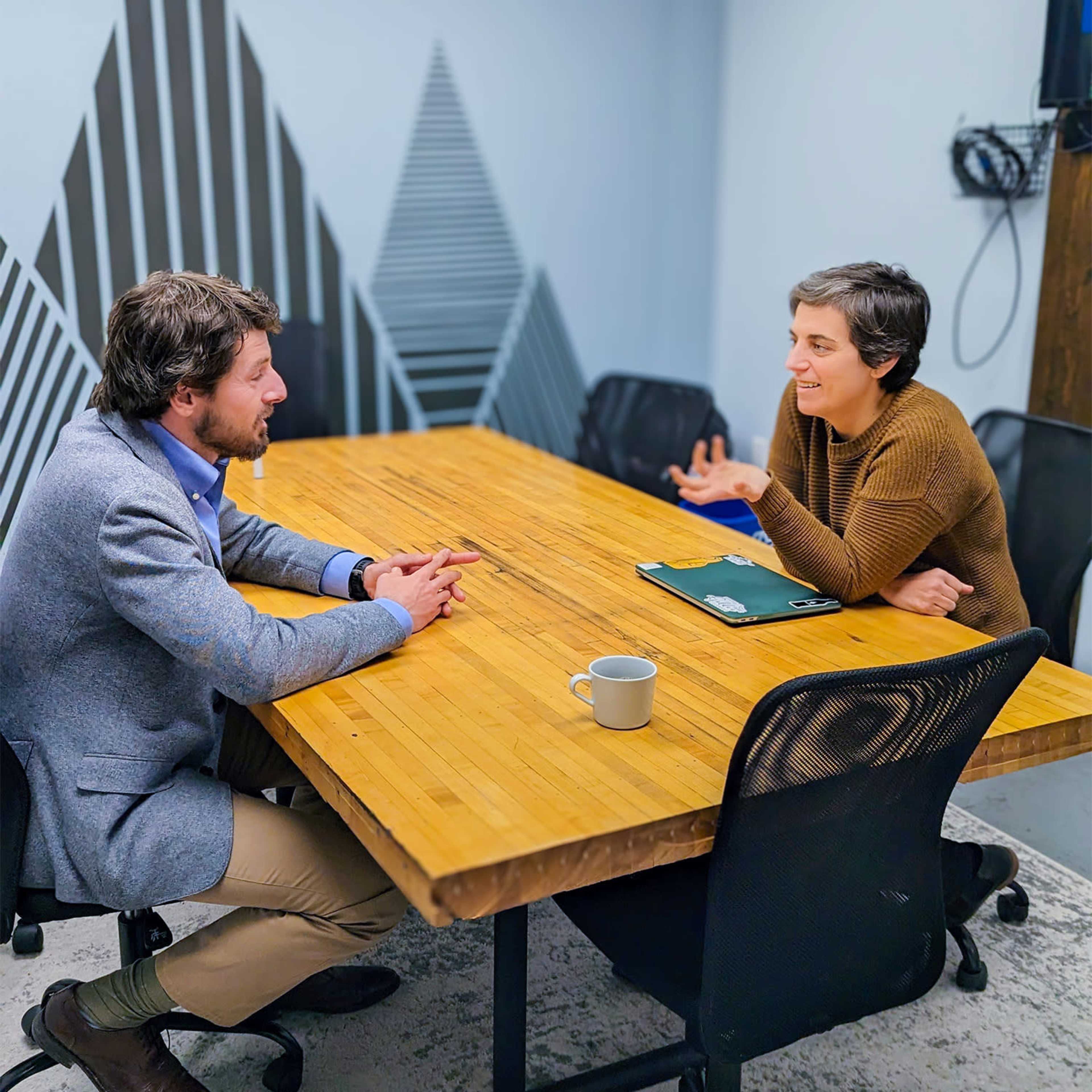 A man and a woman are engaged in conversation at a wooden conference table in a meeting room.