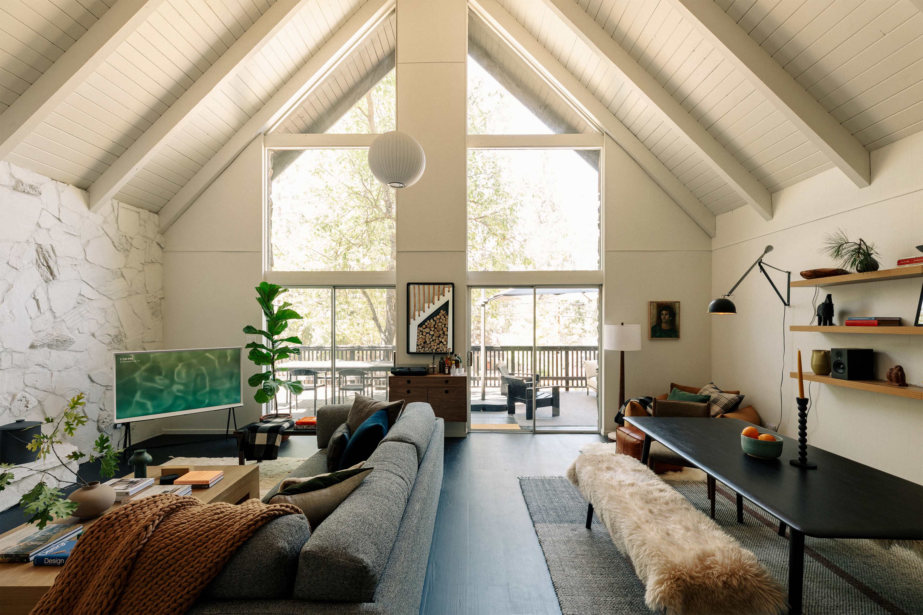 A spacious living room with high ceilings, large windows, a modern sofa, a coffee table, and a television, all complemented by a natural view outside.