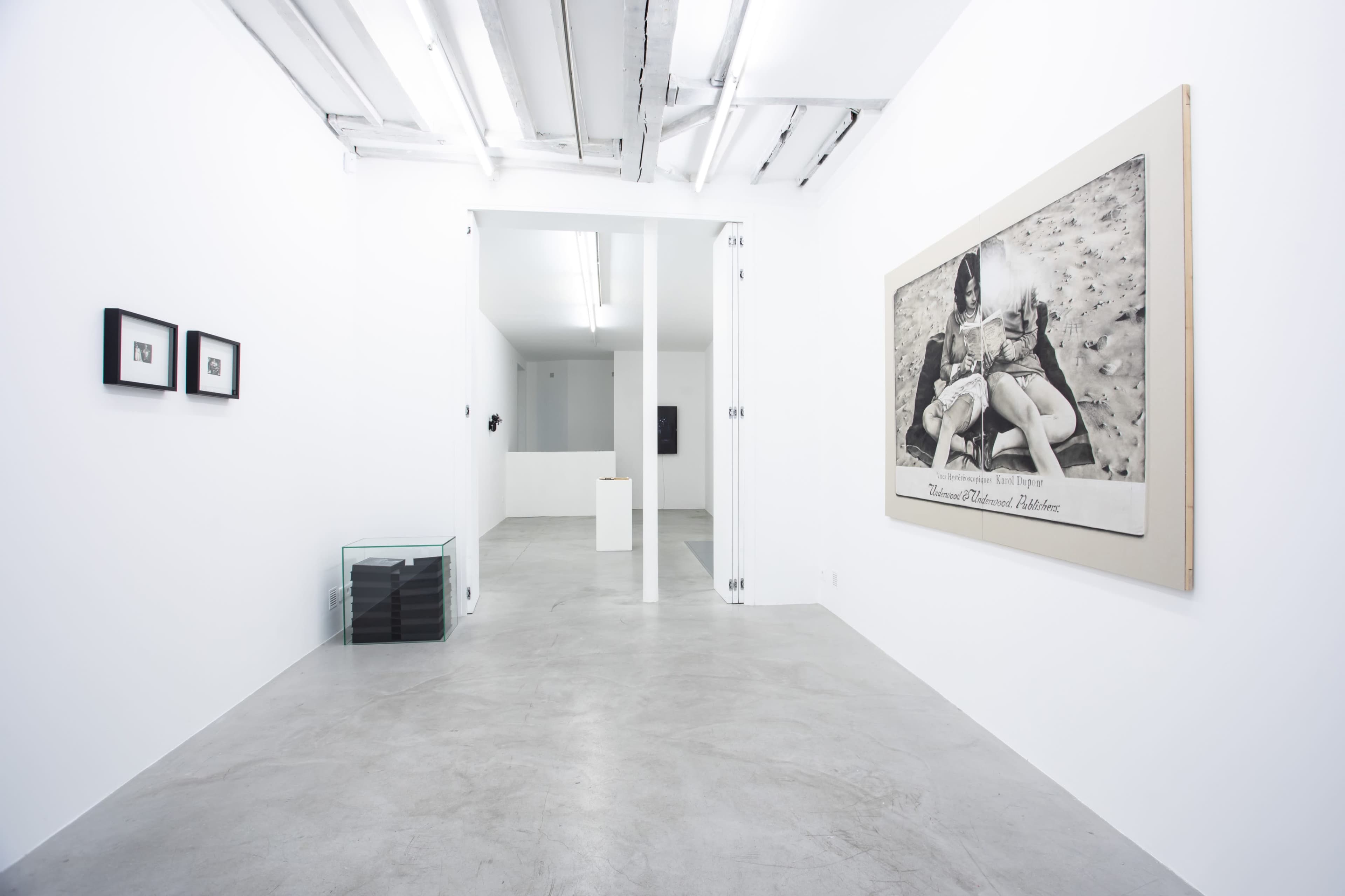 The image shows a minimalist art gallery with white walls, polished concrete floors, and various artworks displayed, including a large framed photograph and smaller pieces on adjacent walls.