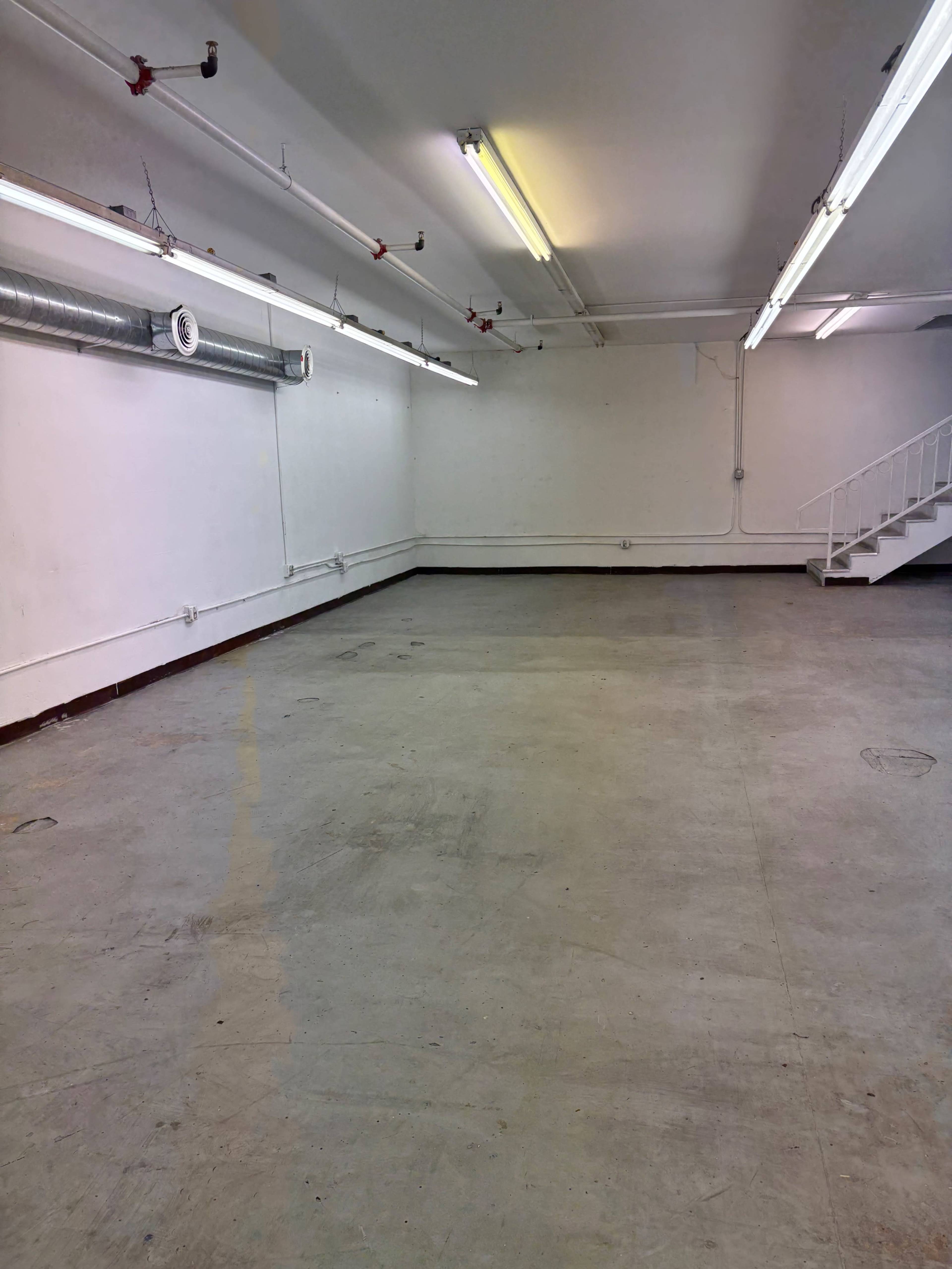 The image shows a spacious, empty room with bare concrete floors and industrial lighting fixtures above.