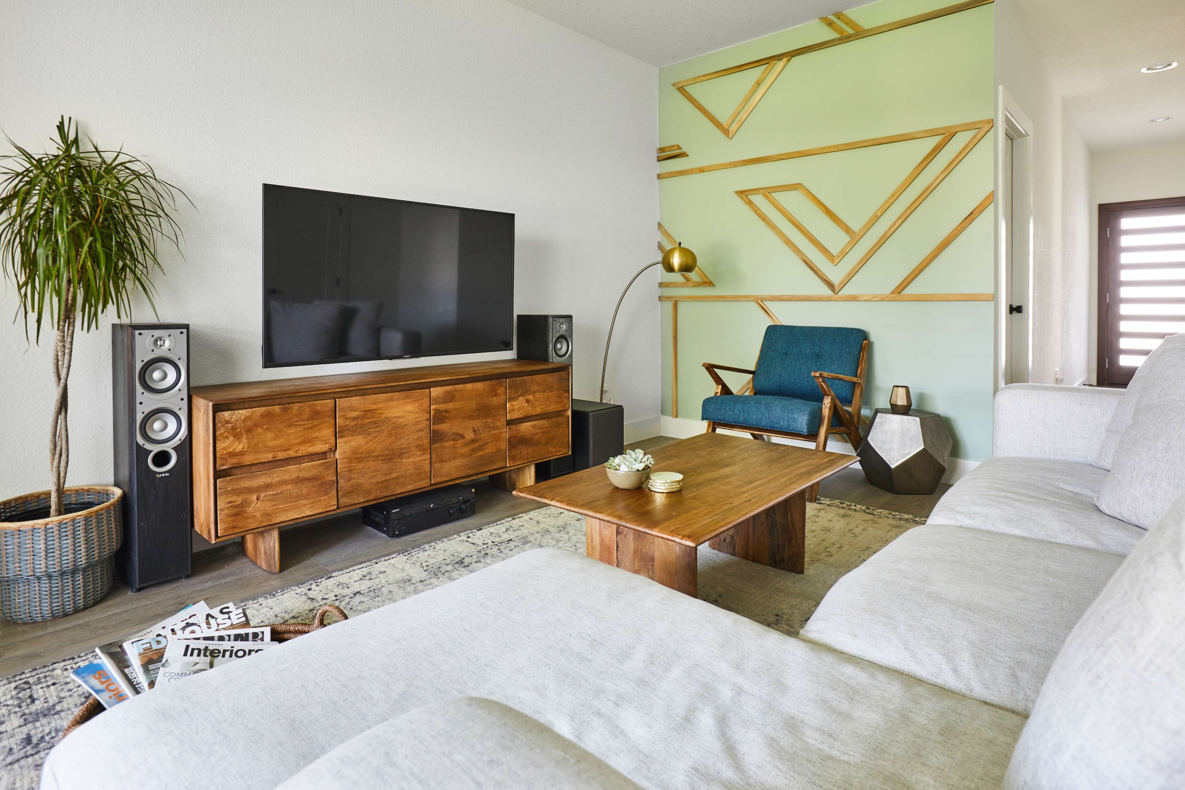 A modern living room features a light-colored sofa, a wooden coffee table, a TV mounted on the wall, and a chair next to a decorative plant.