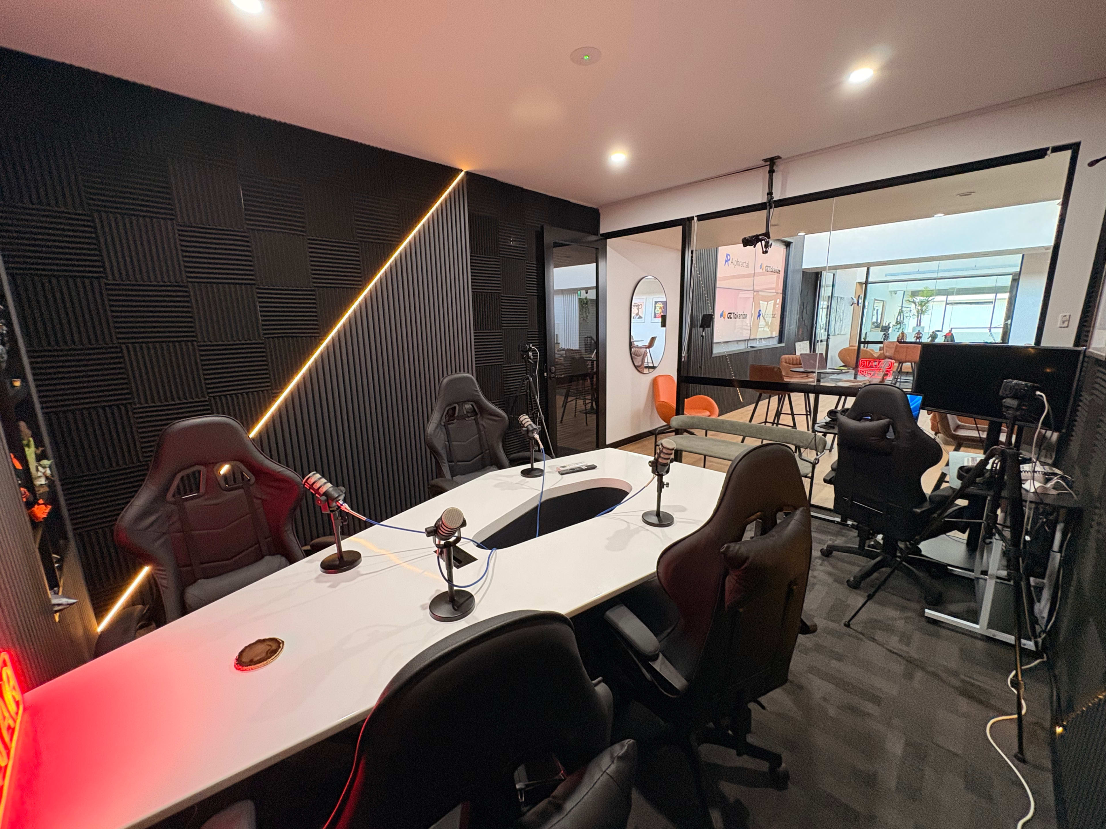 The image shows a modern podcast recording studio with a large white table and several black ergonomic chairs, equipped with microphones and a prominent soundproof wall.