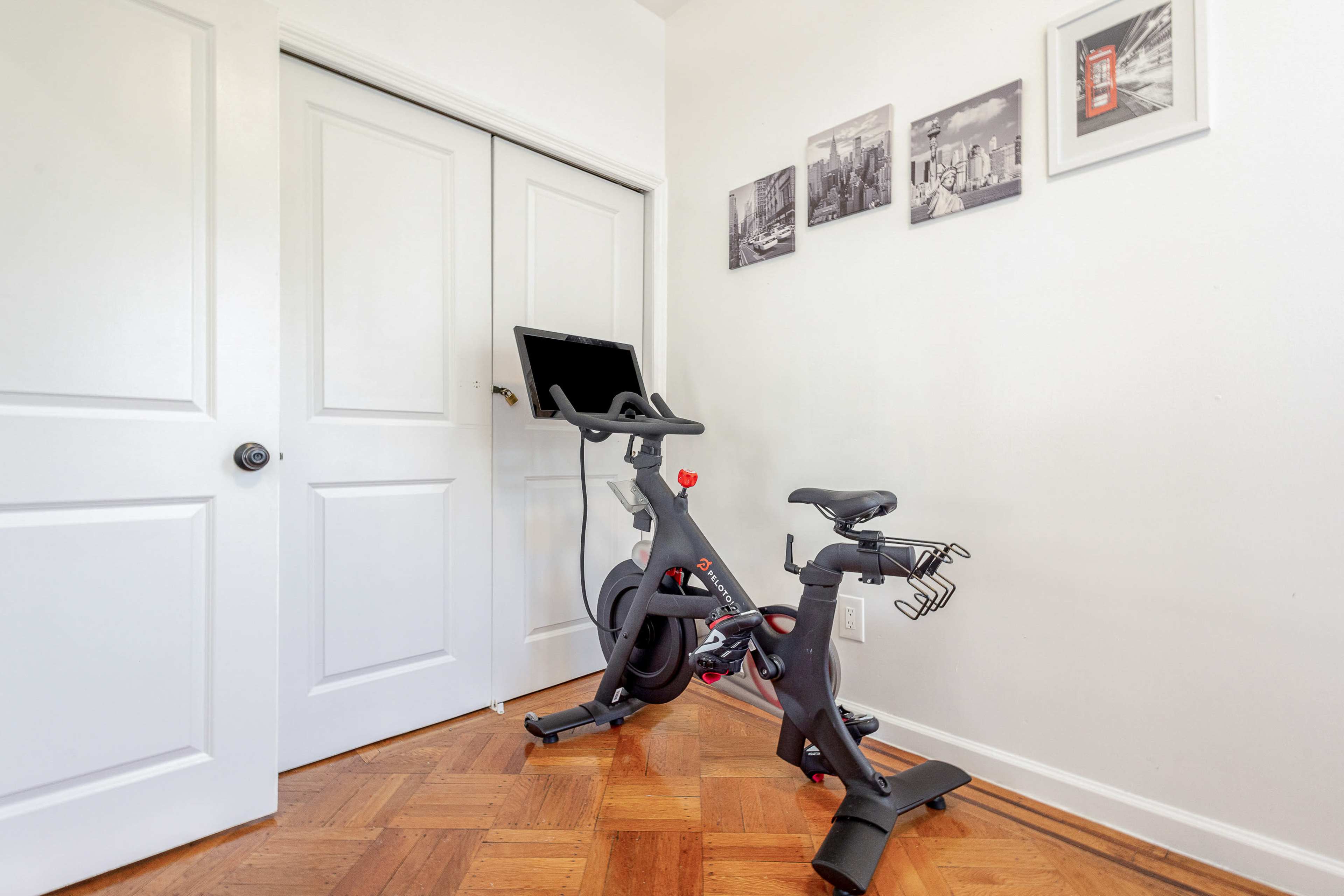 A stationary exercise bike is positioned against a white wall near closed double doors, with framed black-and-white photos hanging above.