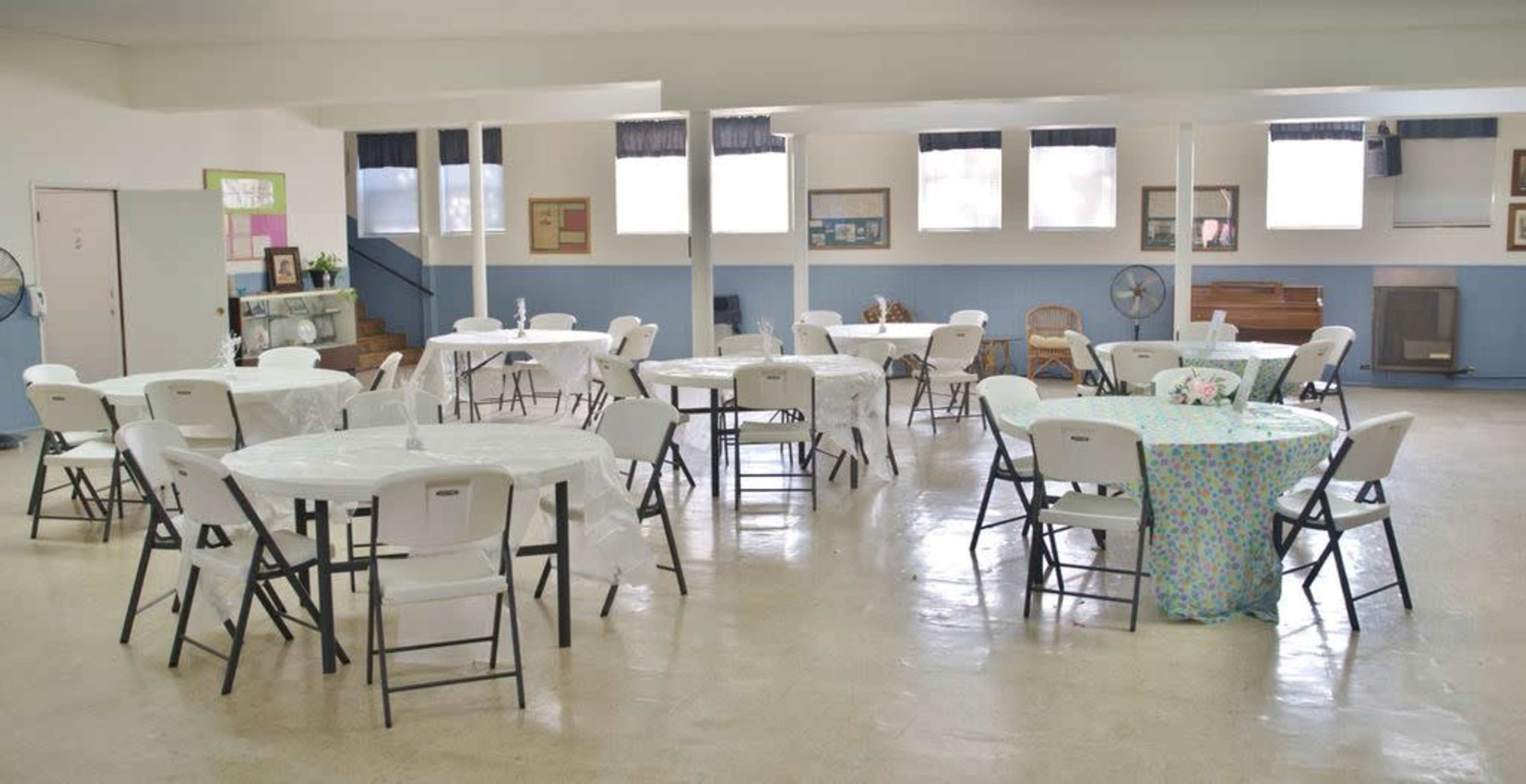 A spacious room is filled with round tables and chairs, some covered with white tablecloths and others with a colorful patterned cloth, set up for an event.