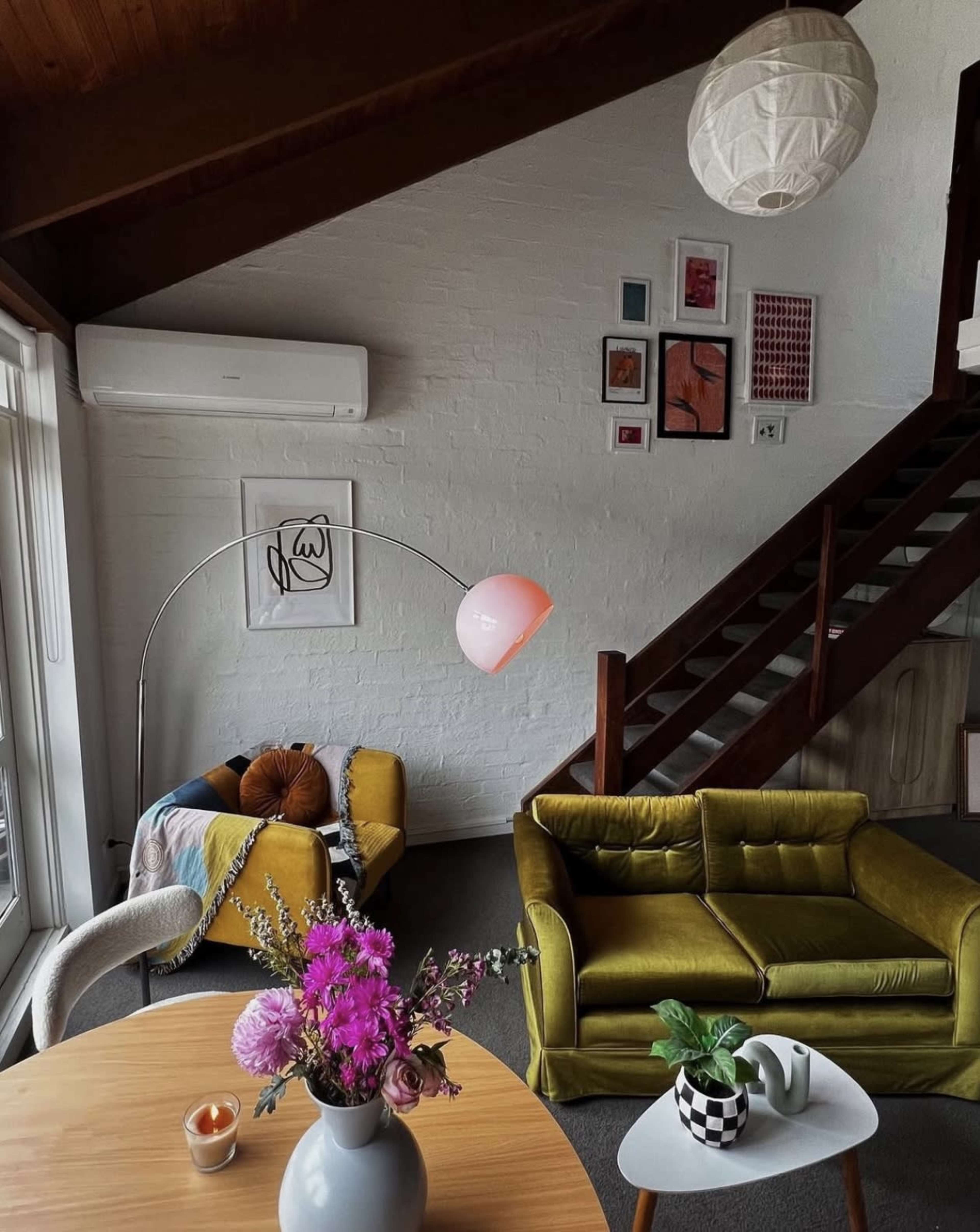 A cozy living room features a yellow couch, a patterned armchair, a round dining table with a vase of flowers, and artwork displayed on the walls.