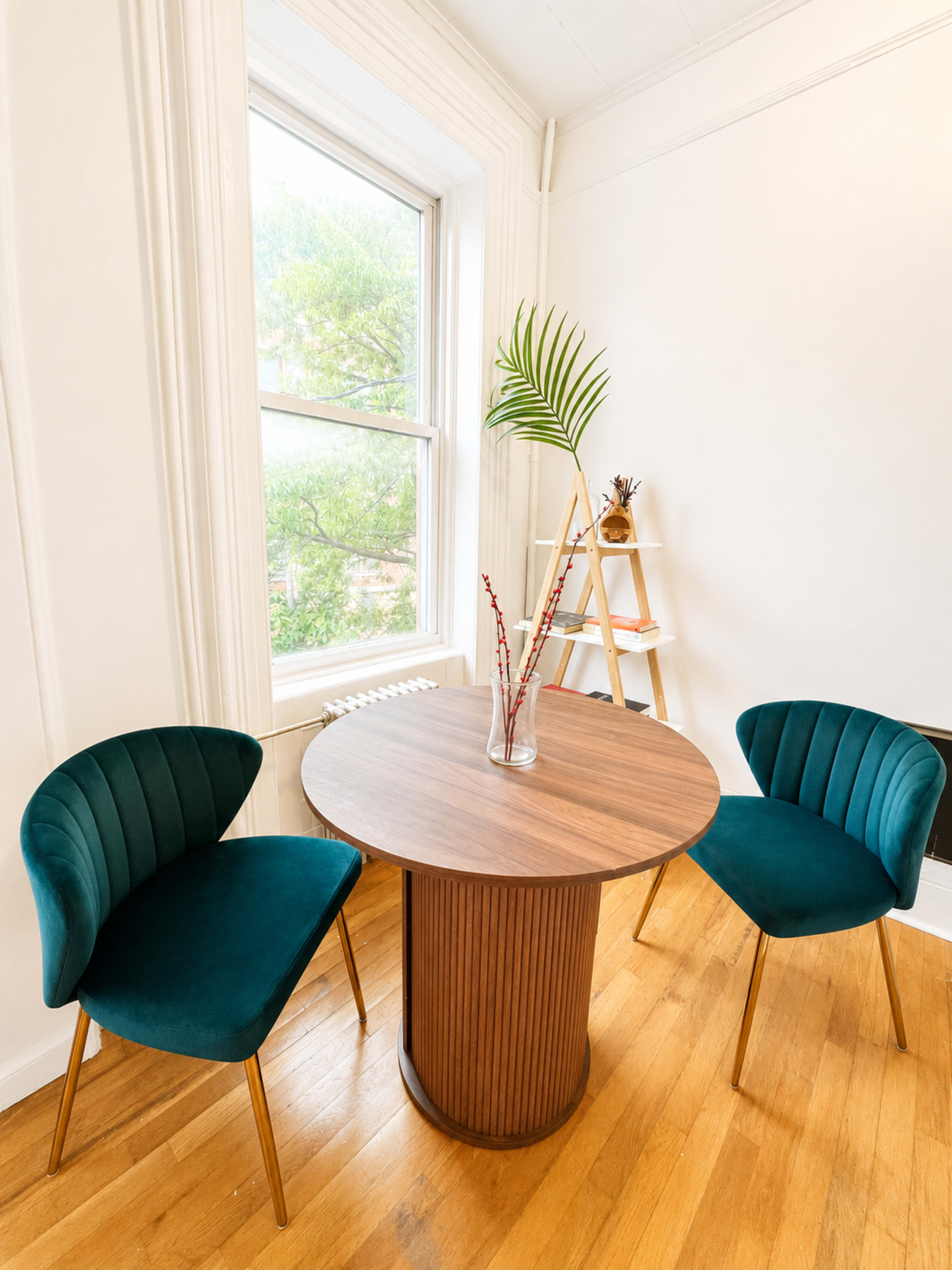 A round table with a wooden base is placed between two teal upholstered chairs in a bright room with a large window.