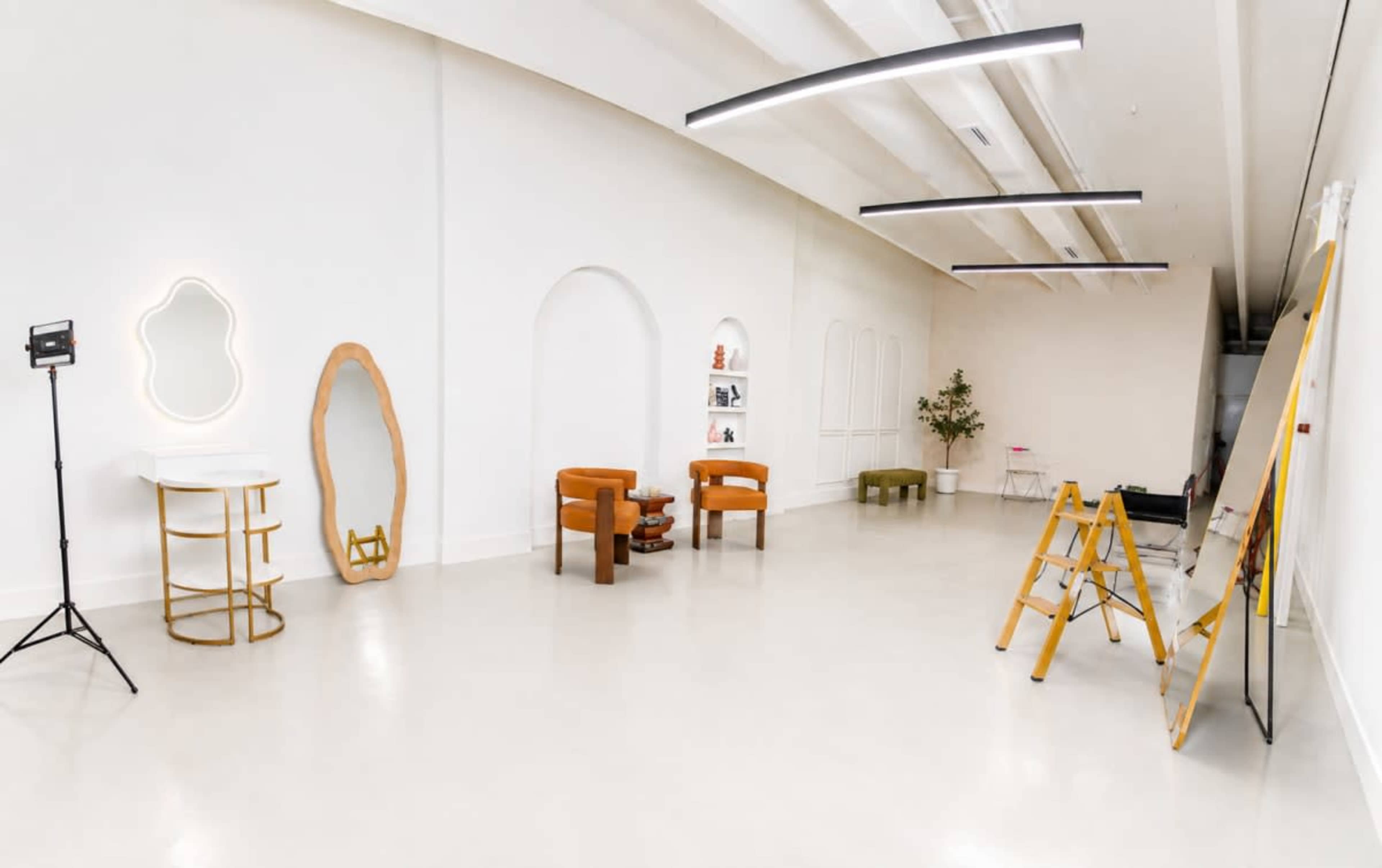 The image shows a spacious, well-lit room with white walls, featuring two orange armchairs, a modern mirror, a small tree, and a couple of furniture pieces along with easels and a lighting fixture.