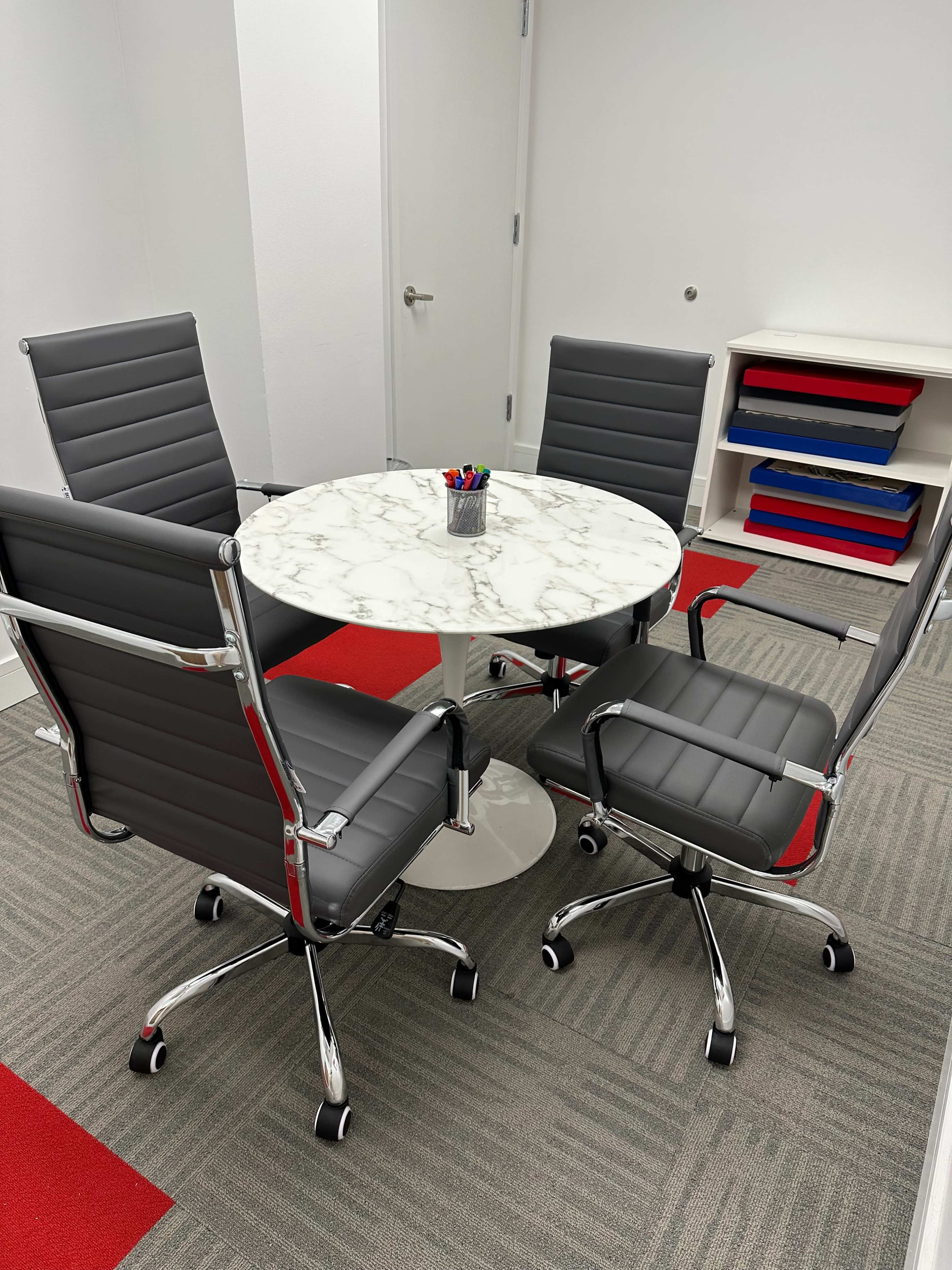 A round marble table with four gray office chairs is positioned on a carpeted floor in a brightly lit meeting area.