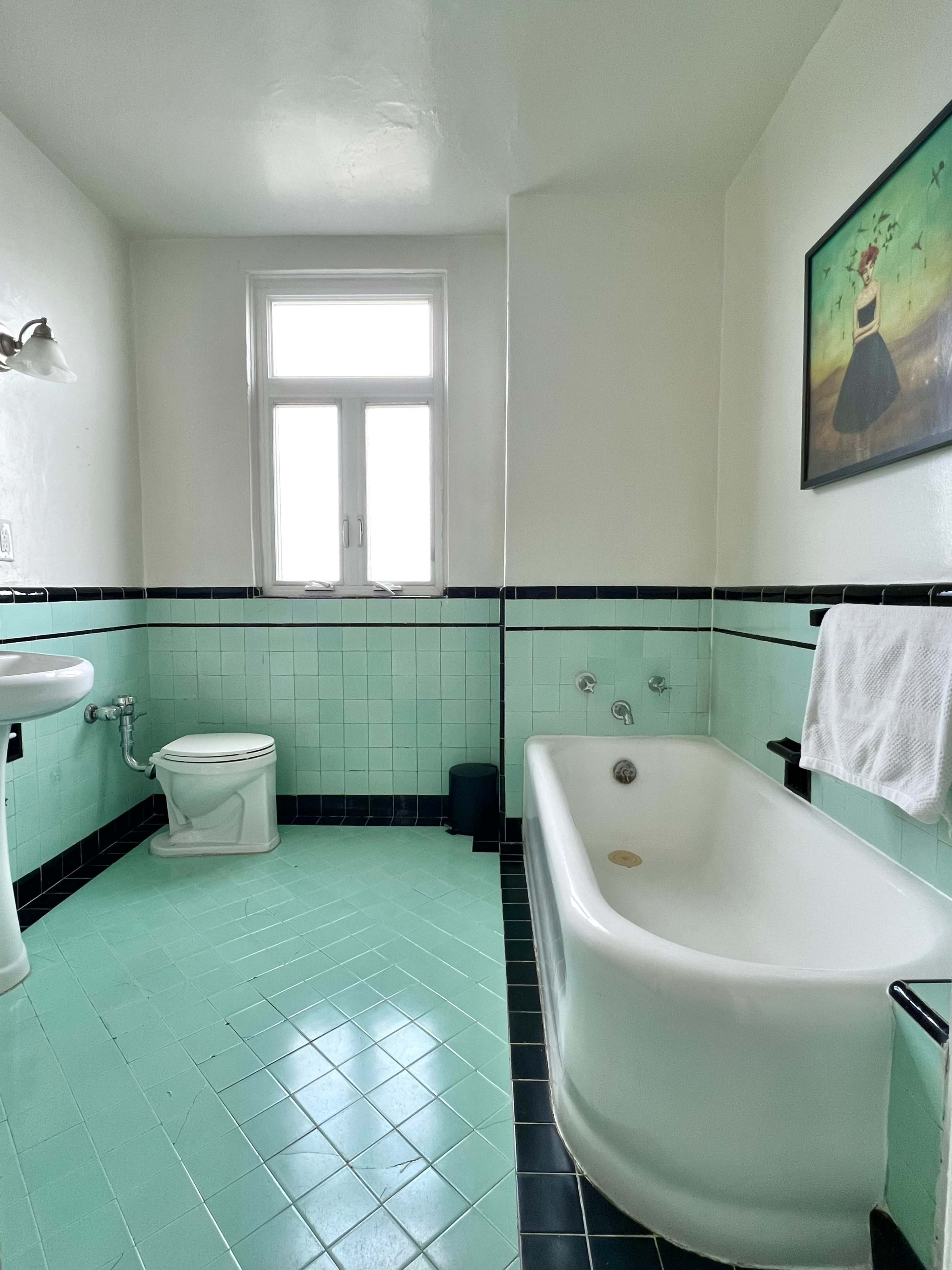 The image shows a bathroom with a turquoise tiled floor, a white bathtub, a pedestal sink, a toilet, and a window allowing natural light.