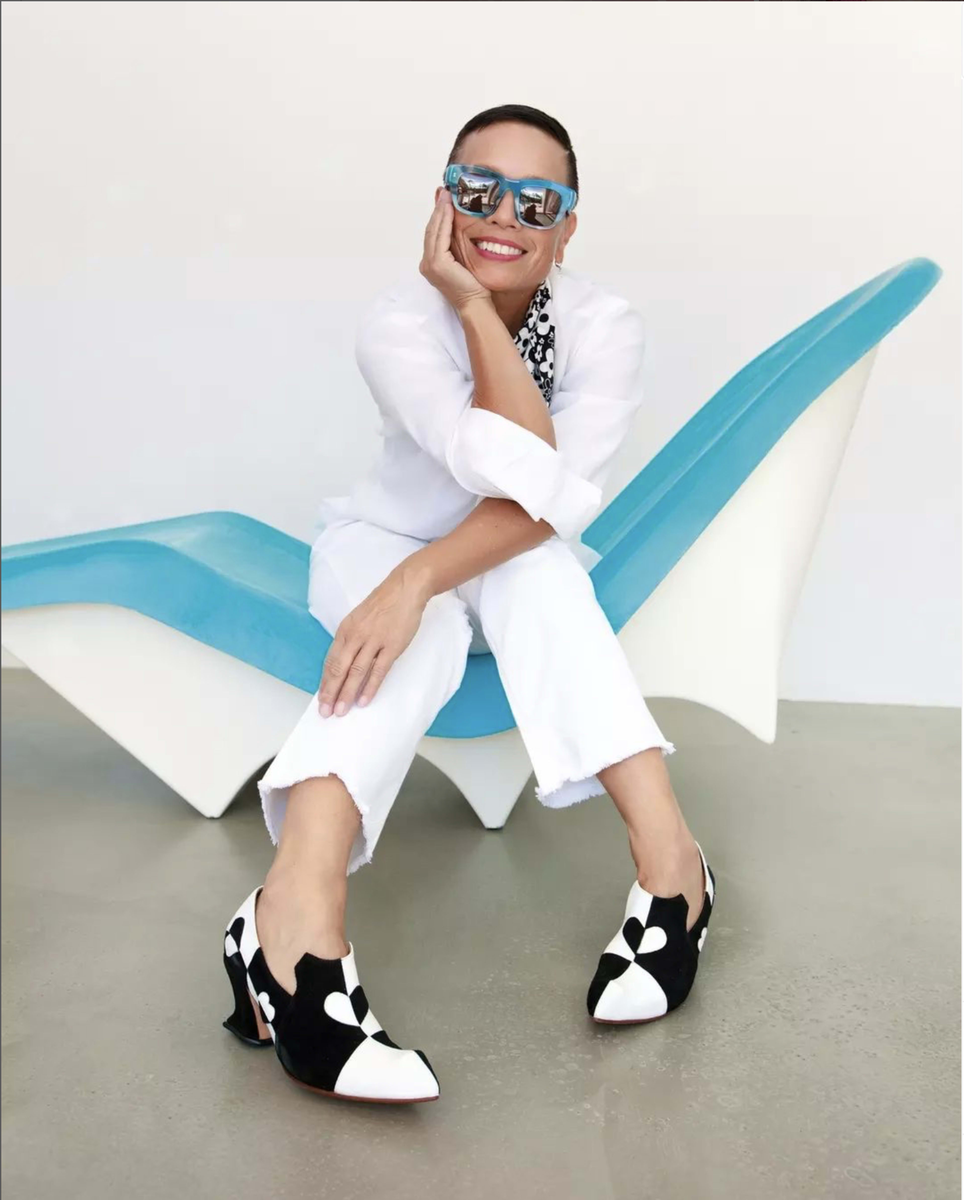A person sits on a blue chair, wearing a white outfit and black and white patterned shoes, smiling while resting one hand on her knee.