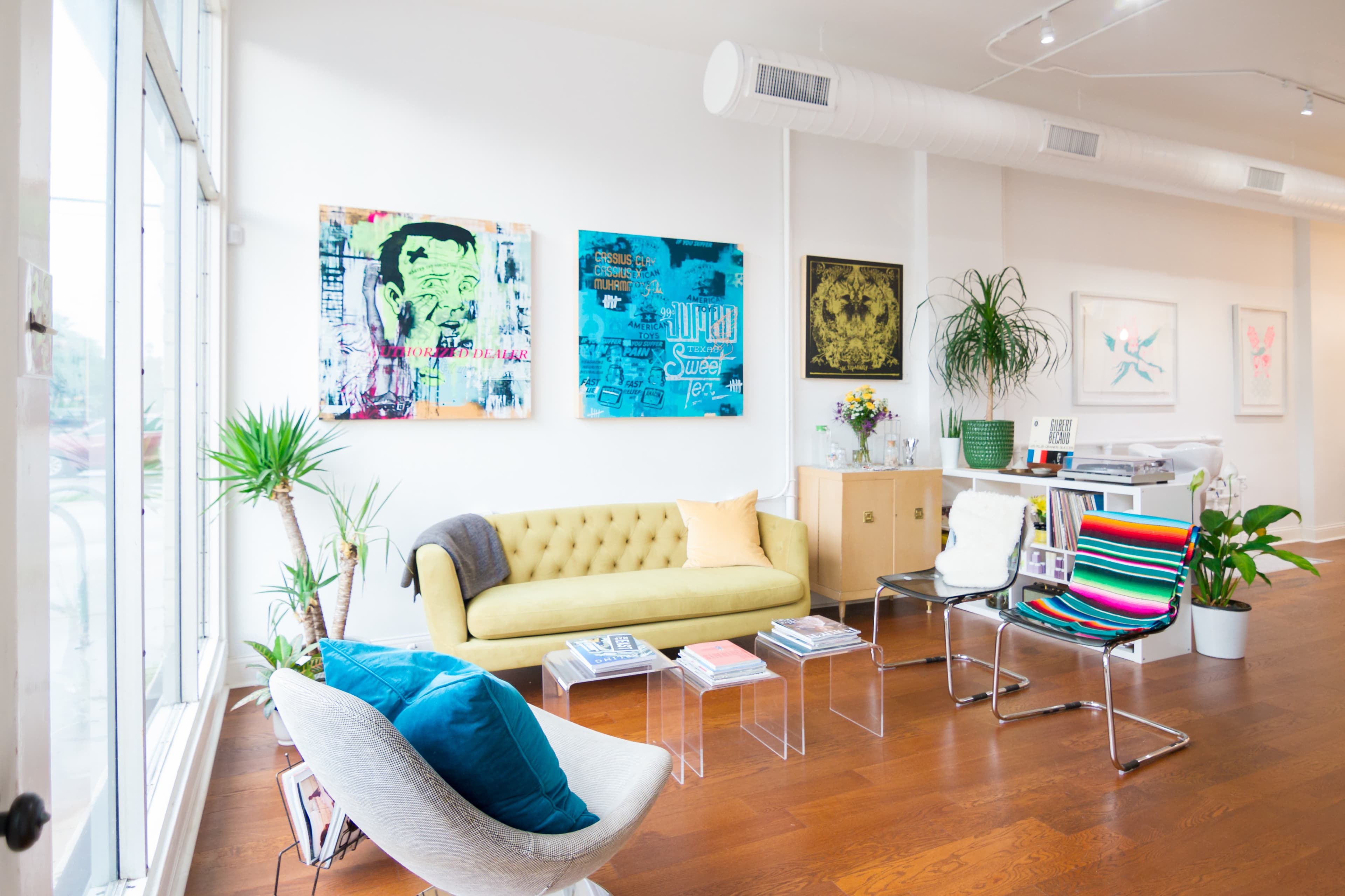 A bright, modern living space featuring a yellow sofa, colorful wall art, a glass table, and various plants.