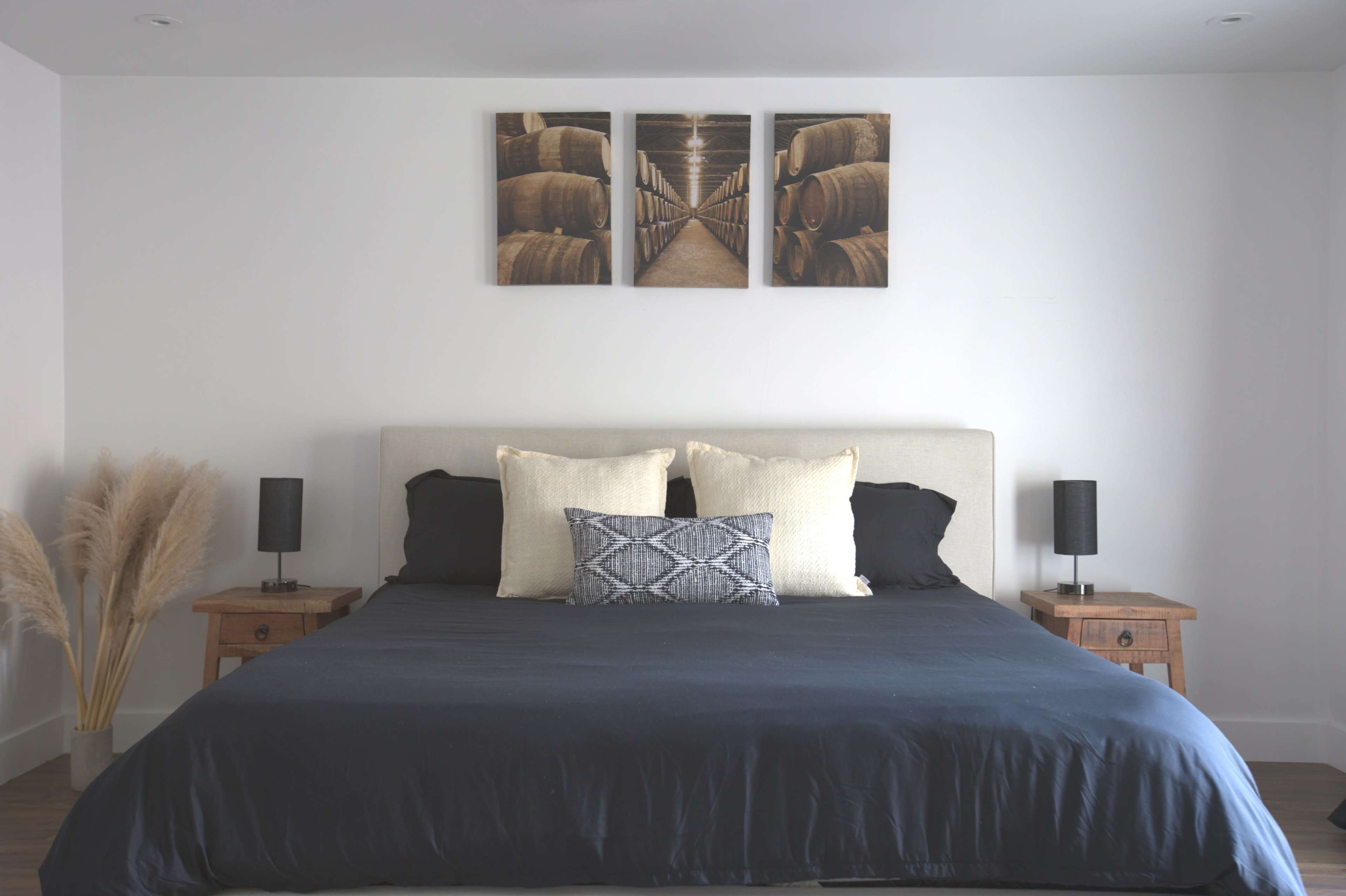 A bedroom with a large bed featuring black bedding and decorative pillows, flanked by two nightstands with lamps, and wall art depicting barrels above the headboard.