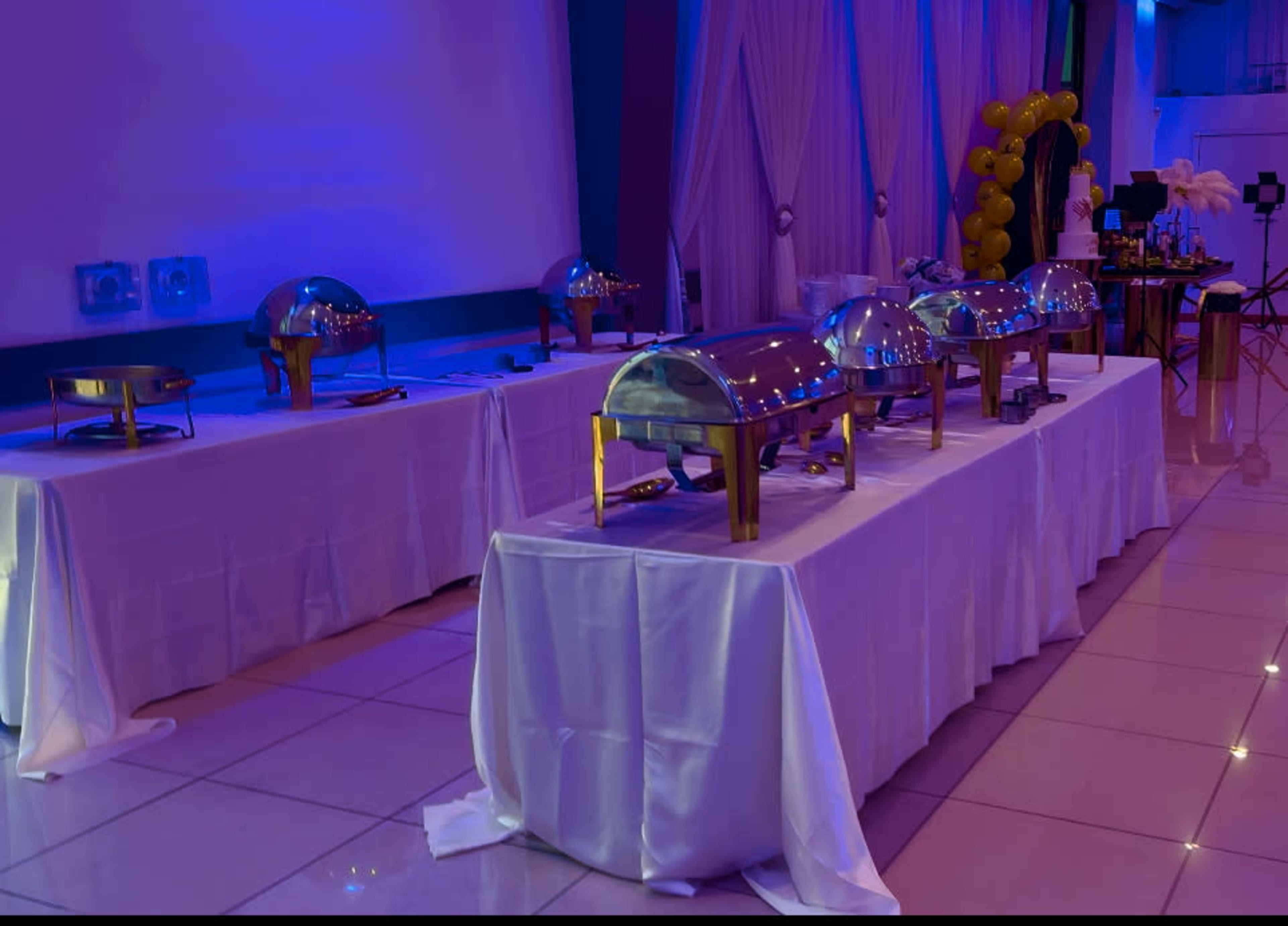 A banquet area features several covered food stations with shiny domes, arranged on white tablecloths under blue lighting.