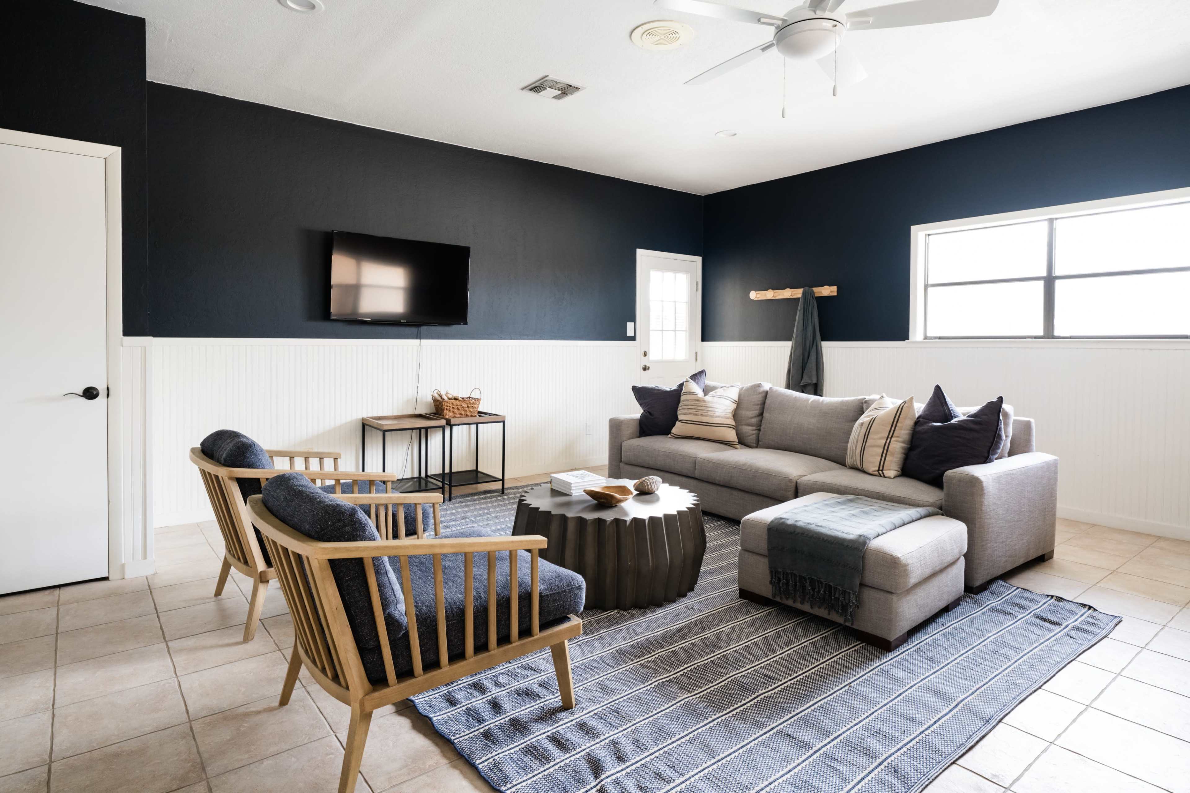 A modern living room features a sectional sofa, two wooden chairs, a round coffee table, and a wall-mounted TV, set against dark walls and a light, tiled floor.