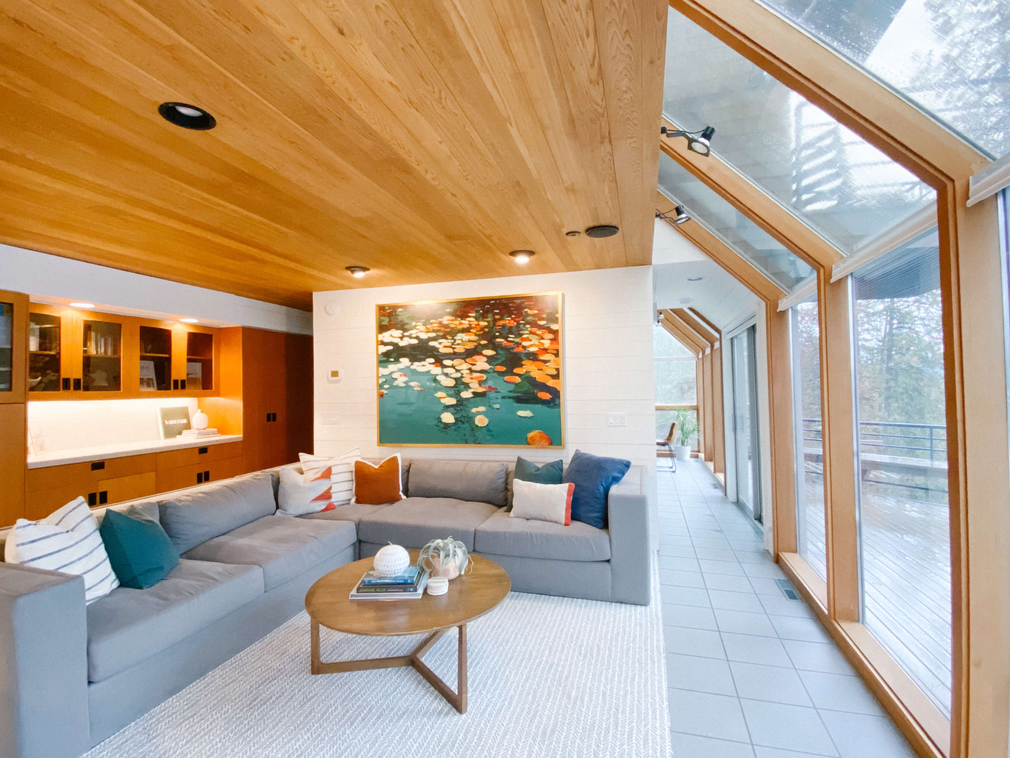 A spacious living room features a sectional sofa, a coffee table, and a large painting, with floor-to-ceiling windows that provide a view of the outdoors.