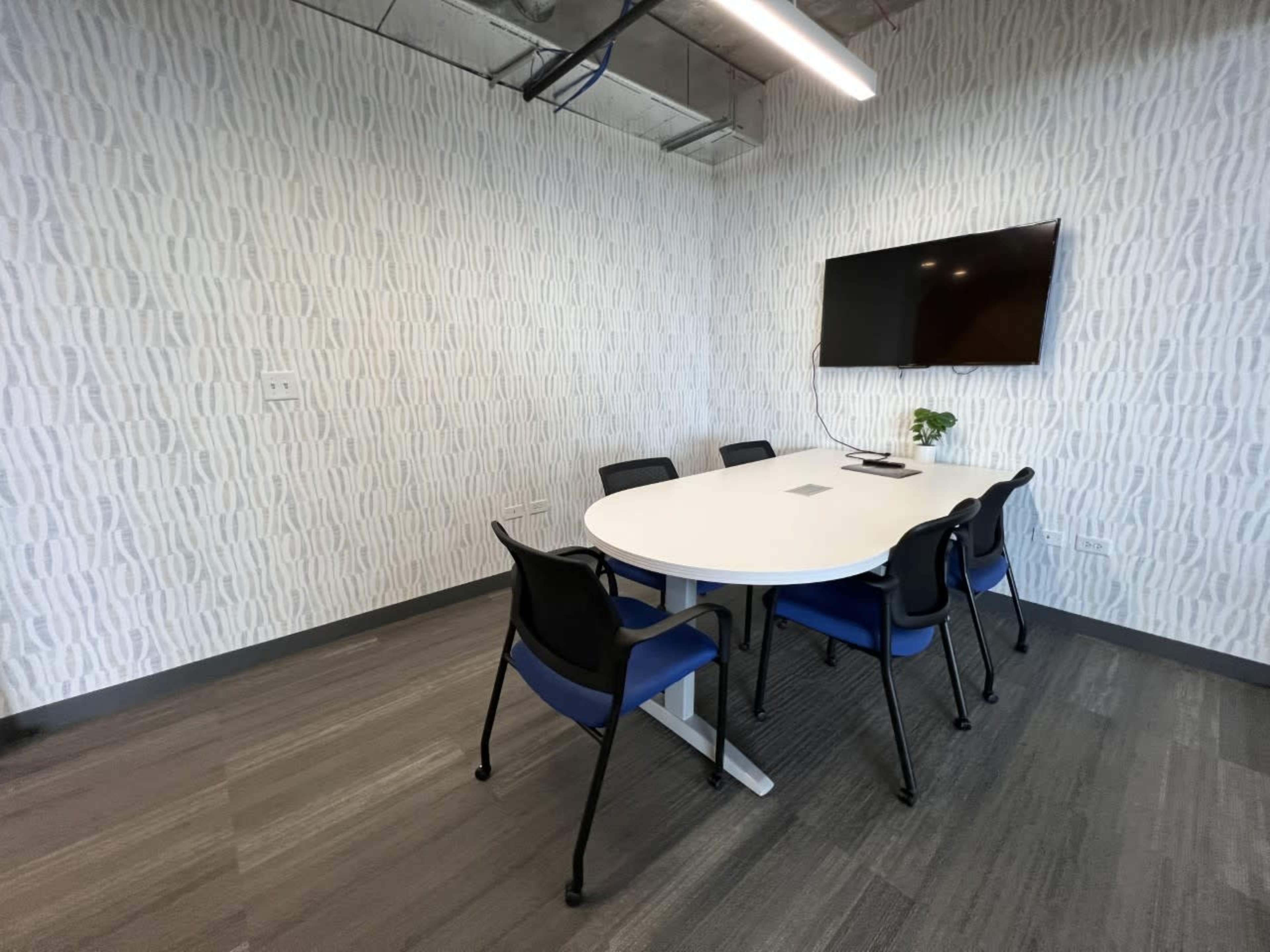 A modern conference room features a round table surrounded by six black and blue chairs, with a wall-mounted television and light-colored patterned wallpaper.
