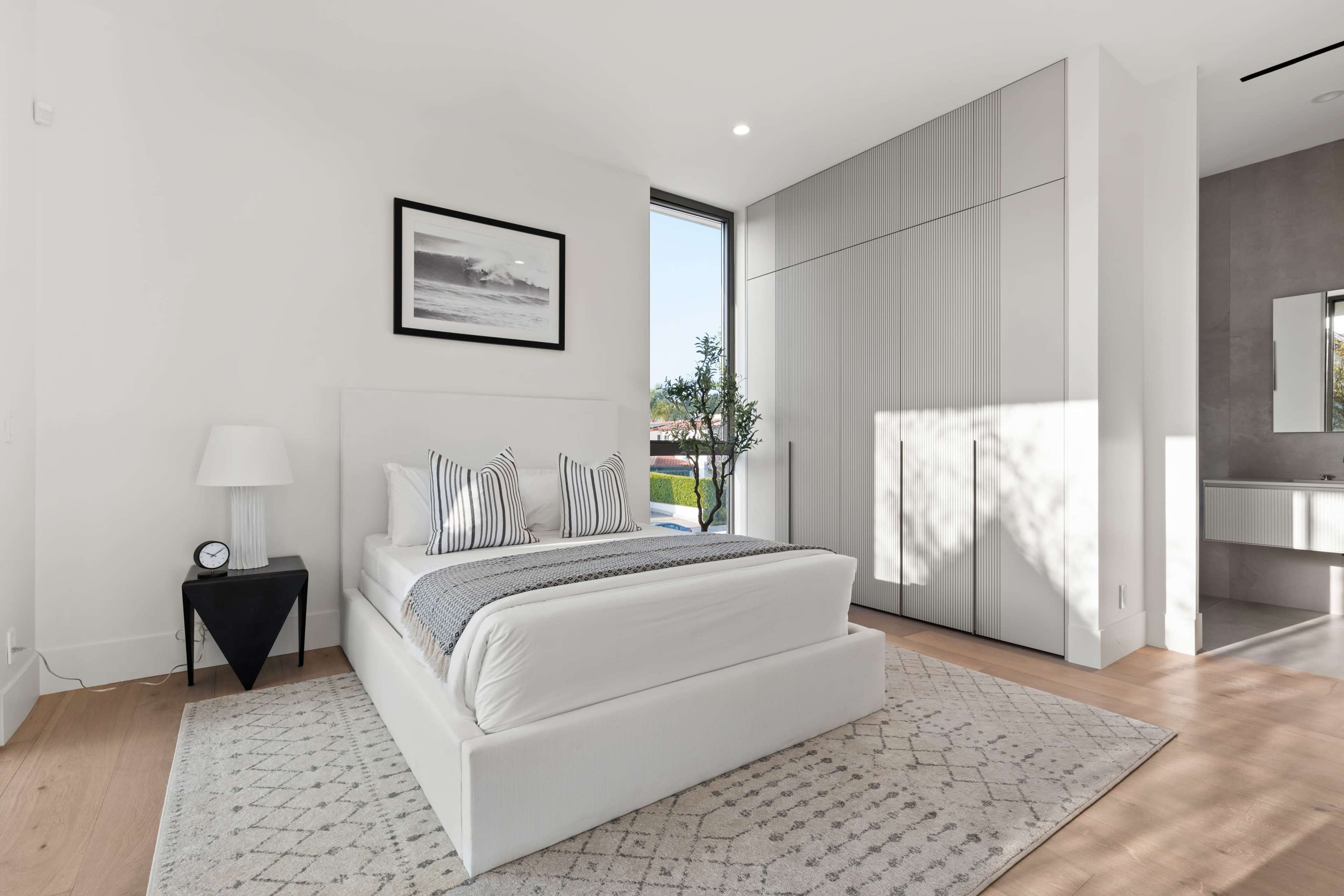 A modern bedroom features a white bed with striped pillows, a bedside table with a clock, and large windows allowing natural light to enter.