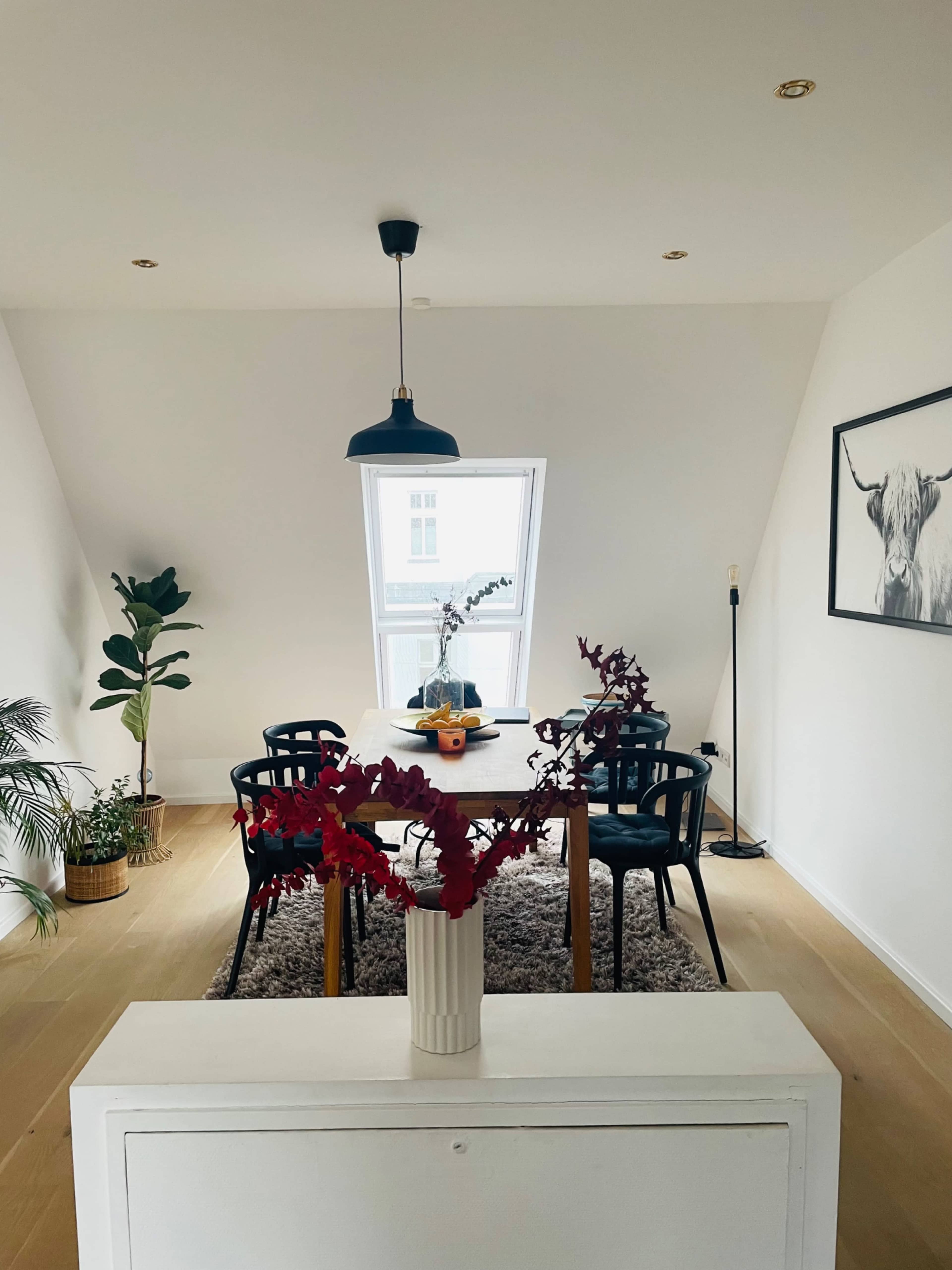 A dining area features a wooden table surrounded by black chairs, with a decorative plant and a vase of red flowers on display.
